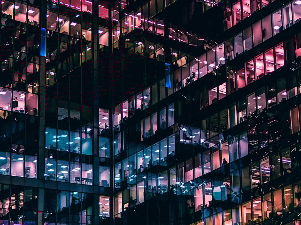 A night view of a modern glass office building, illuminated with colorful lights reflecting the busy interior workspace.
