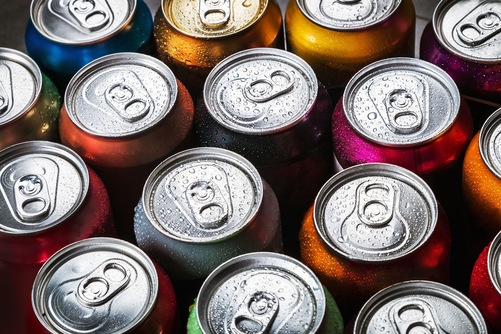 Close-up image of assorted aluminum soda cans with pull tabs, tightly packed together in a top-down view. The cans are in various vibrant colors including red, orange, blue, green, purple, and gold, all covered in condensation droplets, indicating they are cold and freshly chilled.