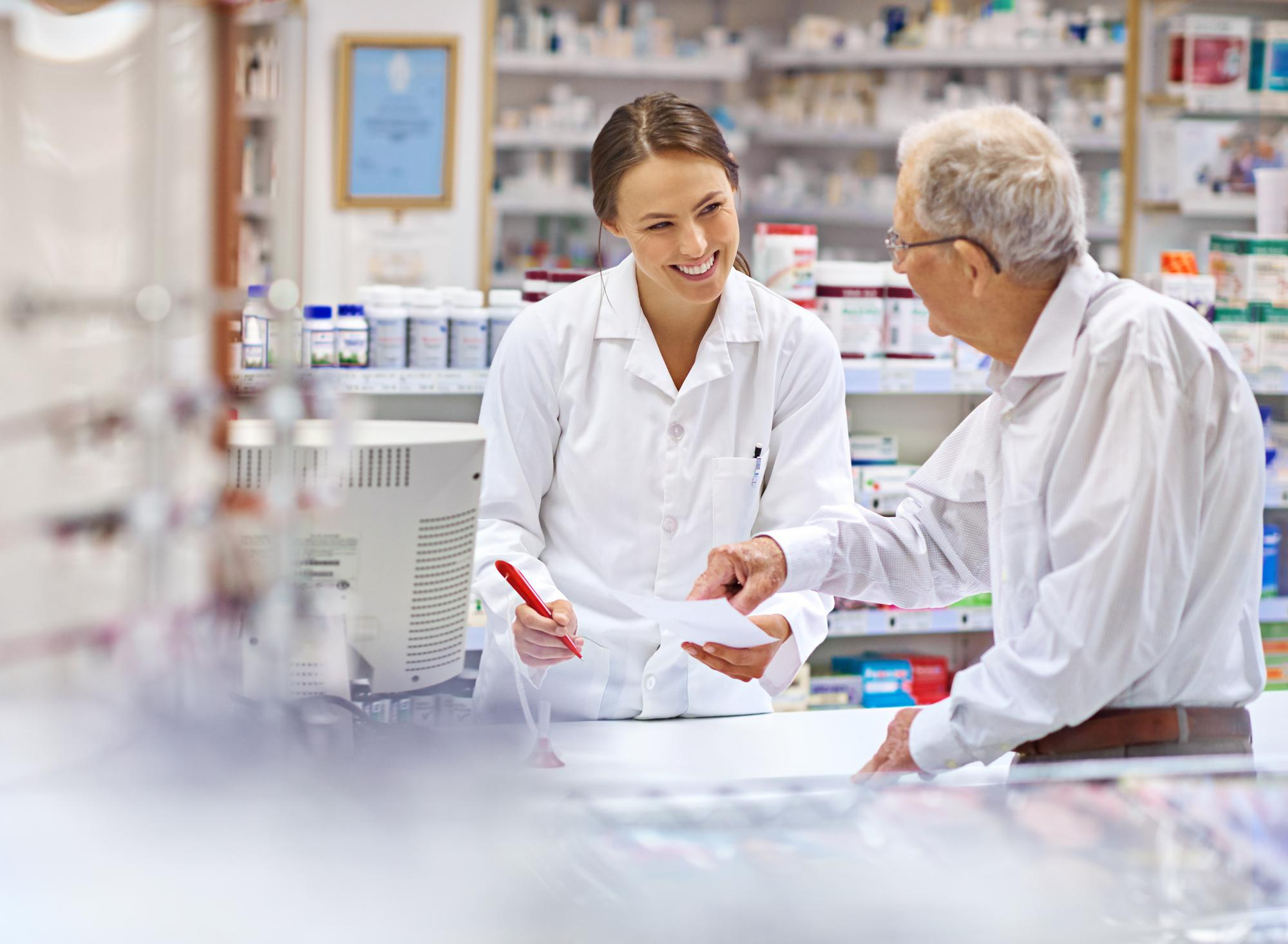 Female pharmacist assisting an elderly male customer with a prescription at a pharmacy counter.