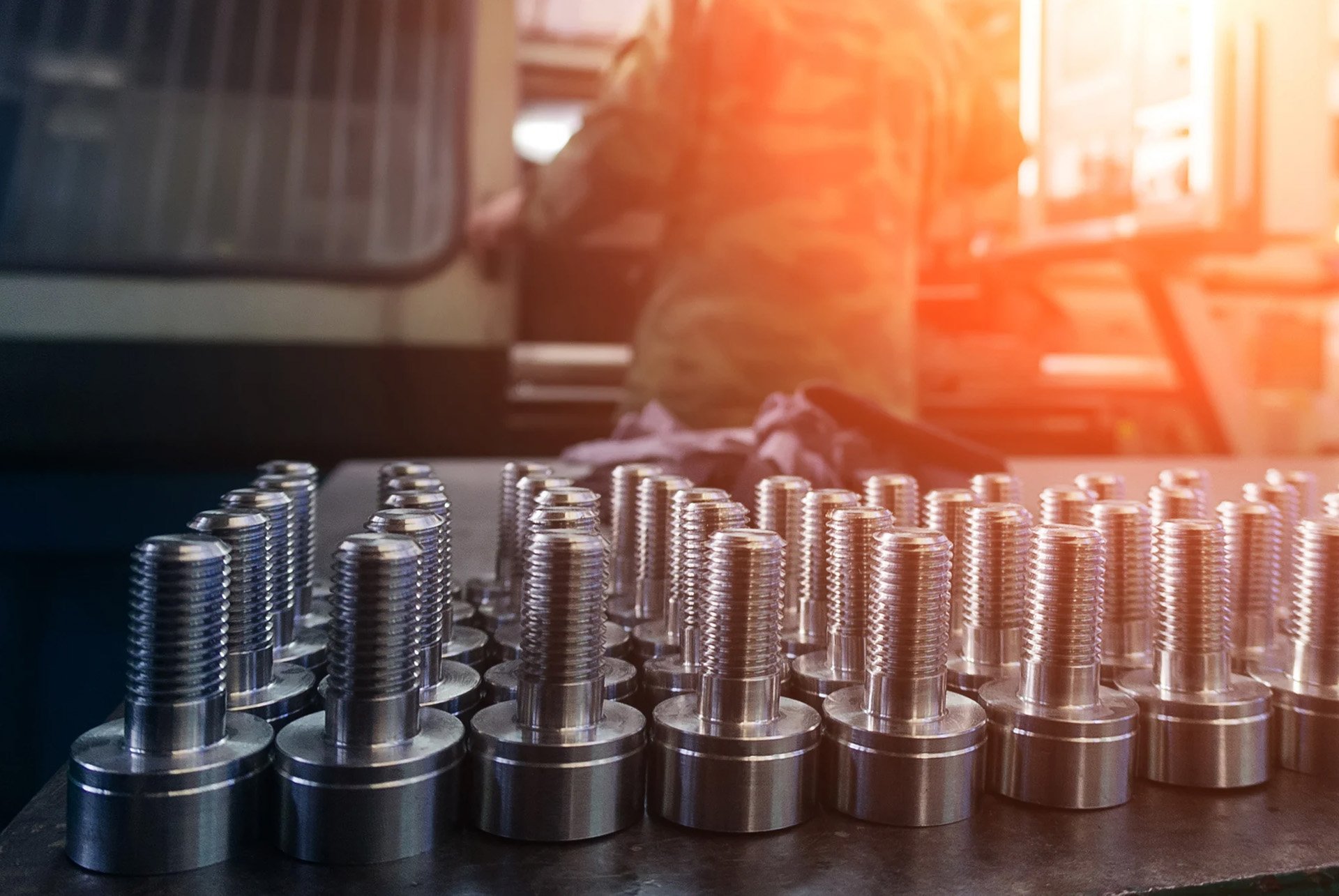 Rows of shiny metal bolts arranged on a table in a manufacturing workshop with a worker in the background.