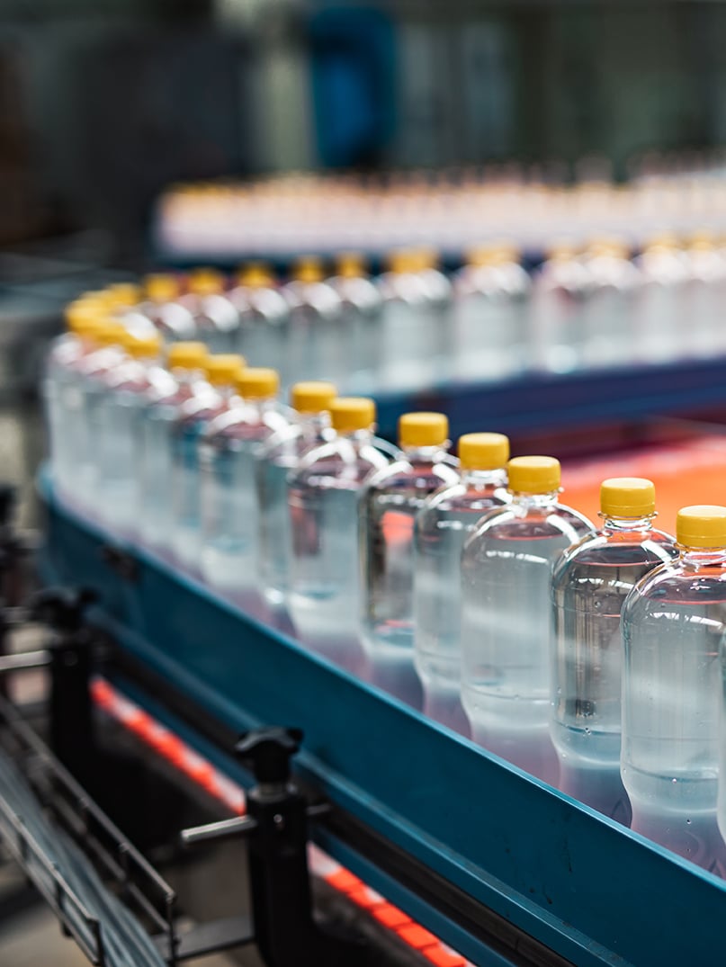 A line of clear plastic water bottles with yellow caps on a conveyor belt in a manufacturing facility.