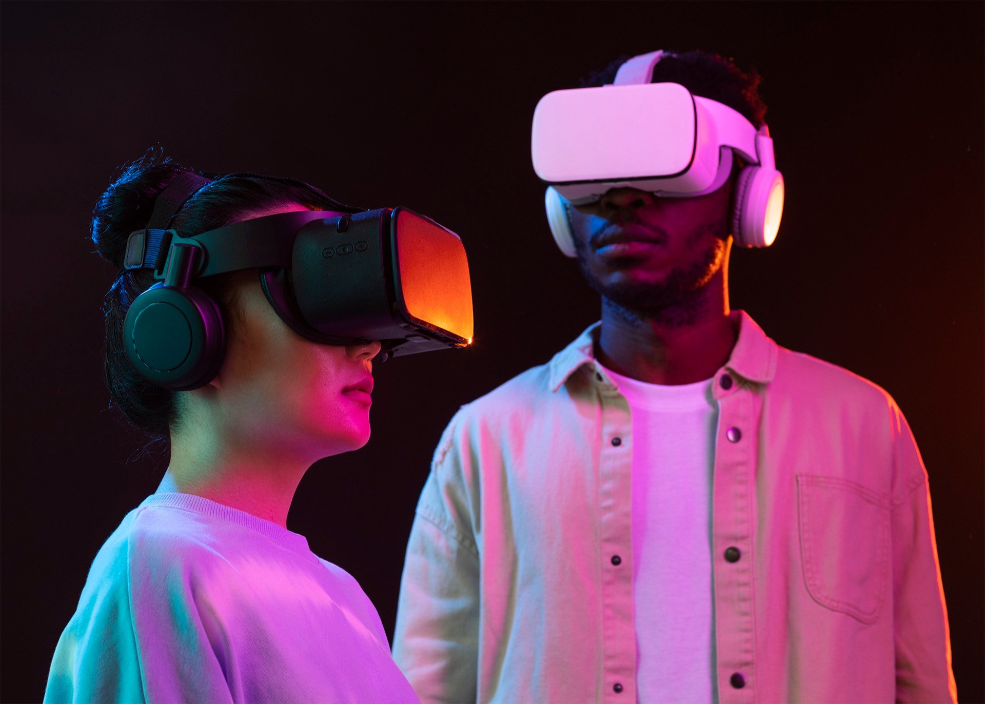 Two people wearing virtual reality headsets and headphones under colorful lighting.
