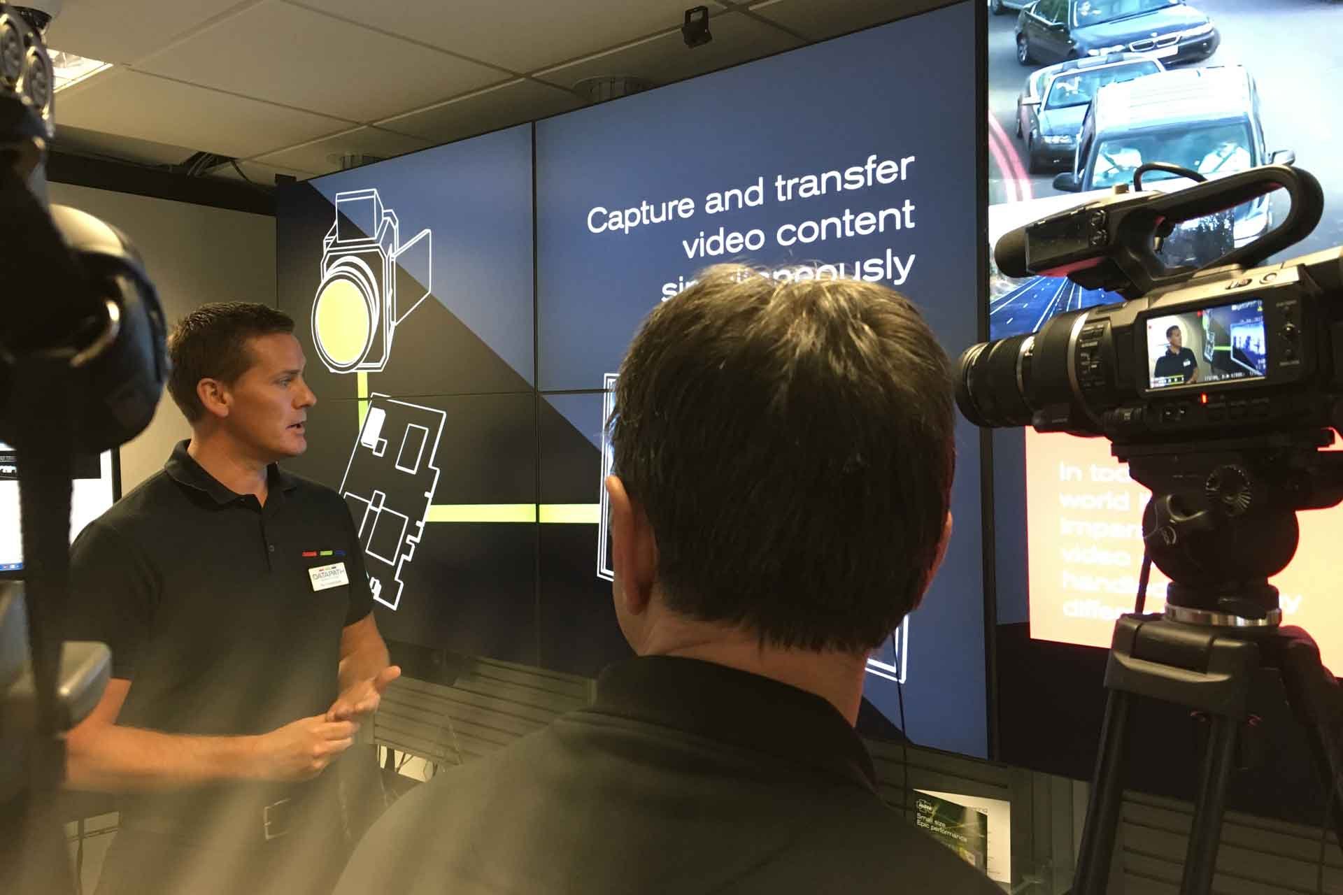 Man speaking in front of a camera with large screens displaying presentation content in the background.