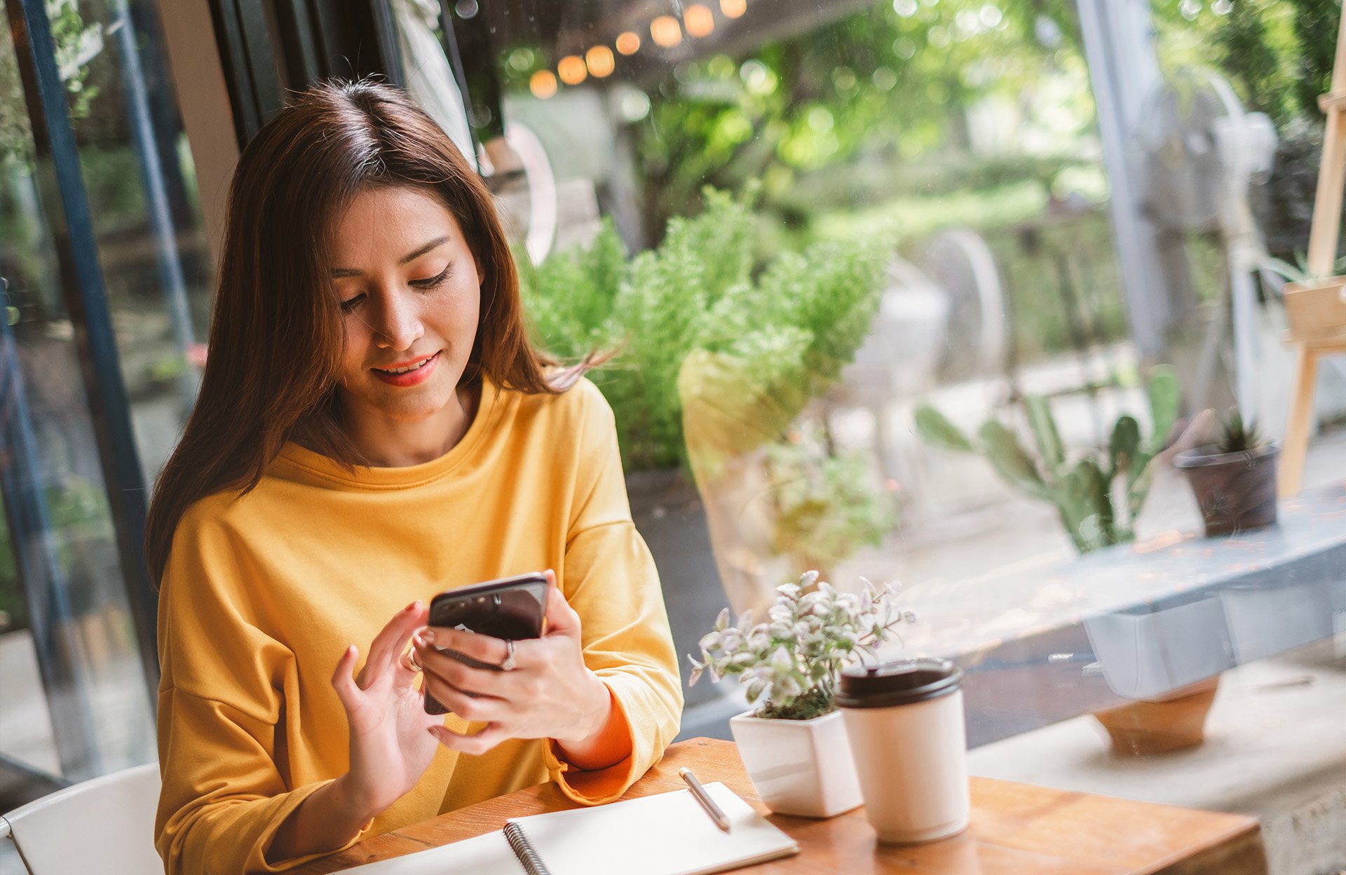 Woman using a smartphone at a cafe with a notebook and coffee cup on the table.