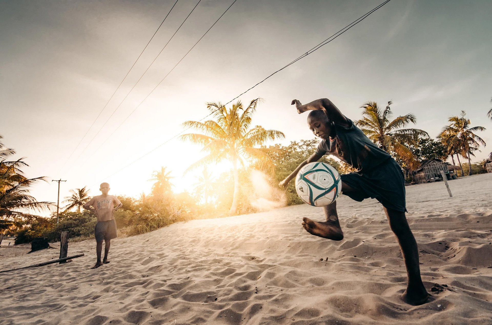 Boy kicking a soccer ball on a sandy beach at sunset with palm trees in the background.
