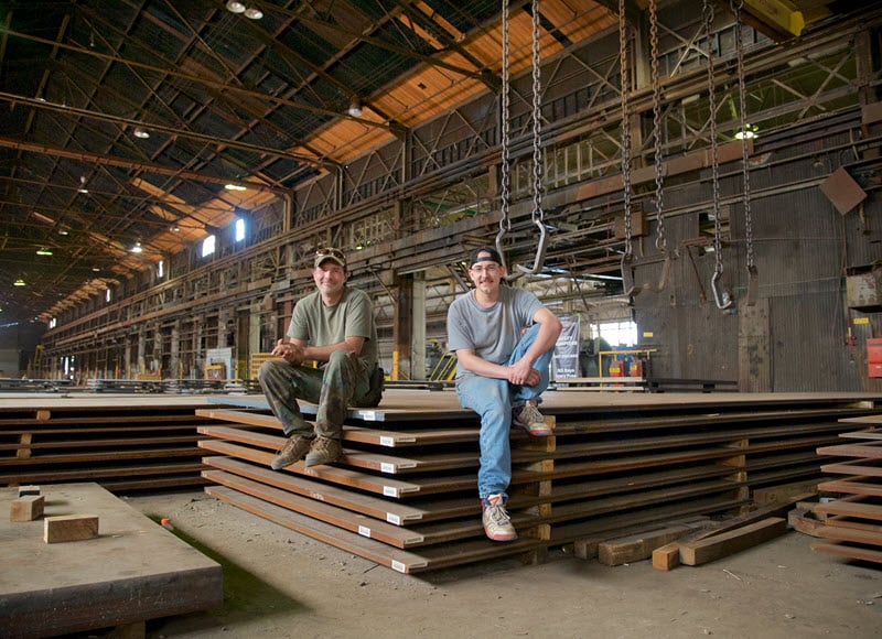 Two workers sitting on stacked metal sheets inside a large industrial warehouse.