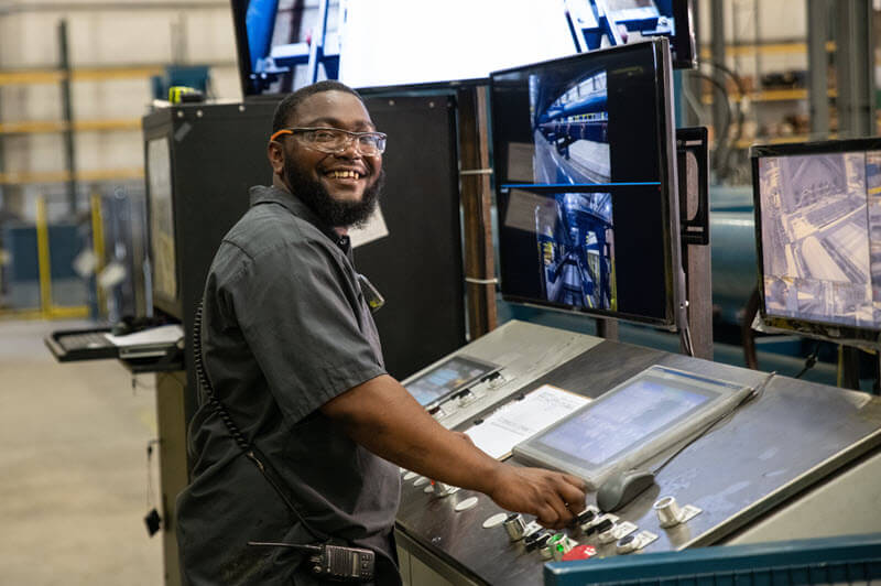 Smiling man operating a control panel with multiple surveillance screens in a factory.