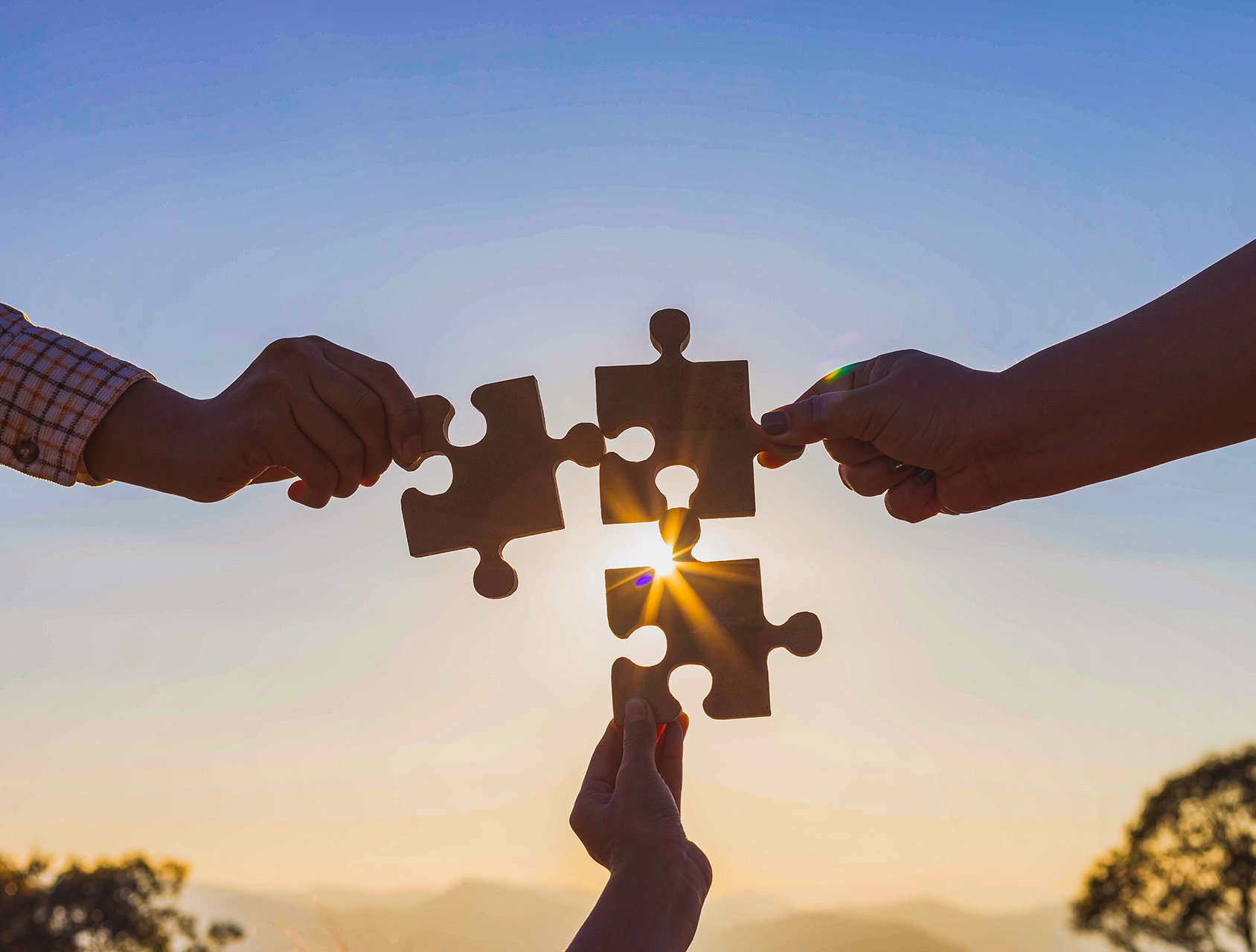 Hands holding wooden puzzle pieces against a sunset backdrop, symbolizing collaboration and connection.