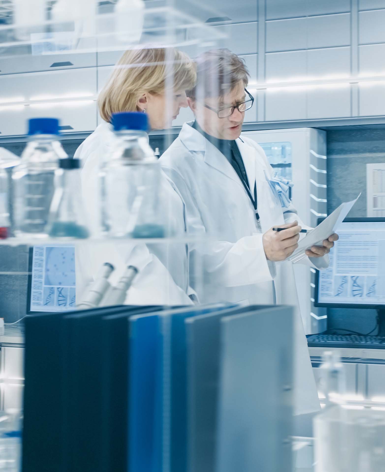 Two scientists in lab coats discuss data in a modern laboratory, surrounded by lab equipment and glass bottles on a shelf.