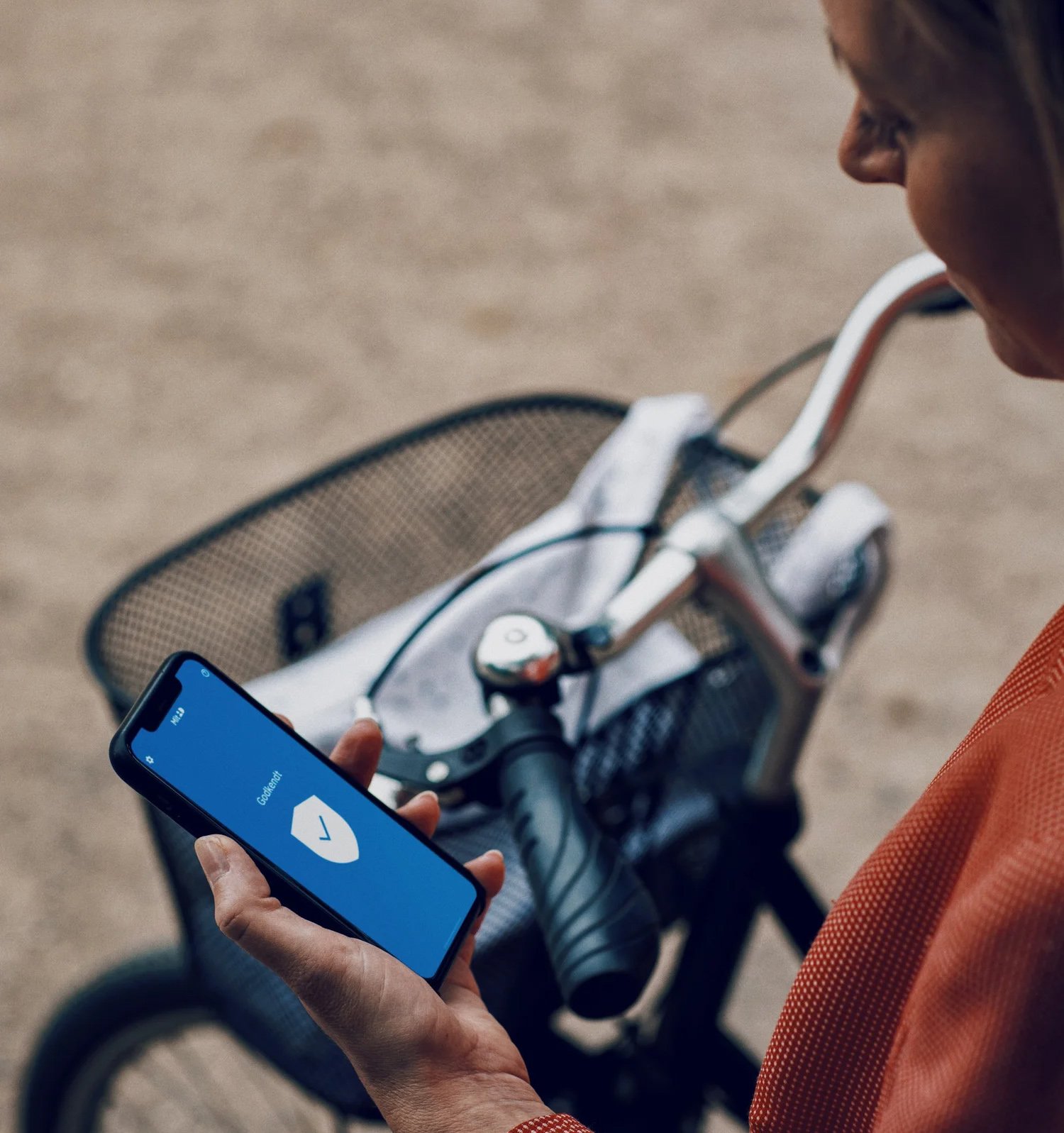 A person holding a smartphone with a blue screen displaying a shield icon, beside a bicycle with a woven basket.