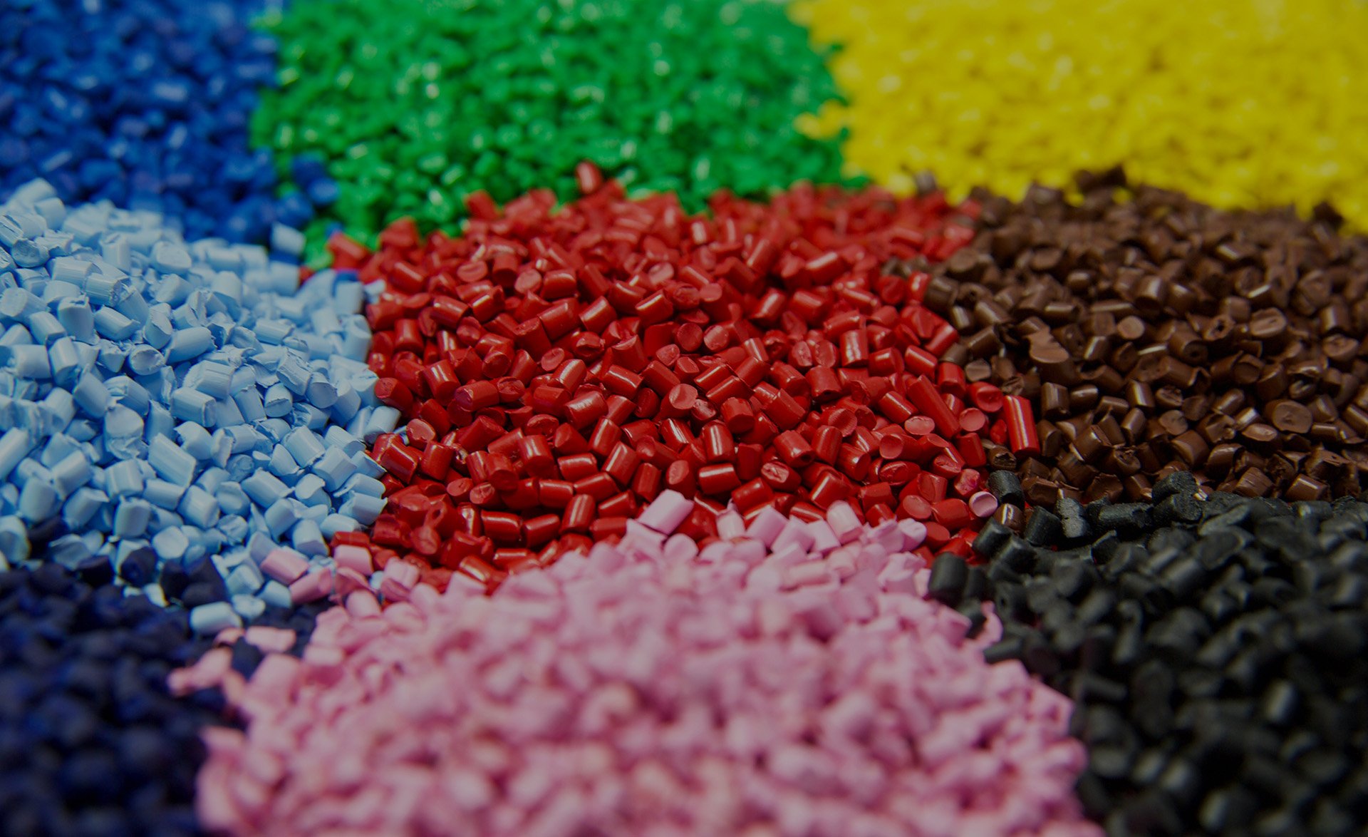 Close-up image of multicolored plastic polymer granules arranged in a grid-like pattern. Each pile contains small cylindrical and cuboid-shaped plastic pellets in vibrant colors including red, green, yellow, black, brown, blue, light blue, pink, and purple.