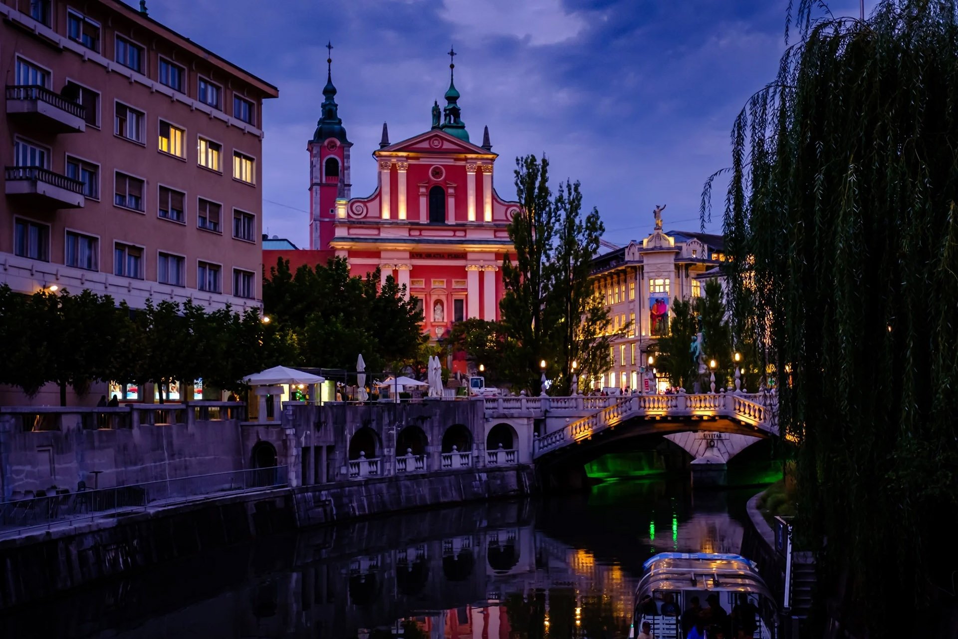 A night view of a city with a river, illuminated buildings, and a bridge reflecting on the water's surface.