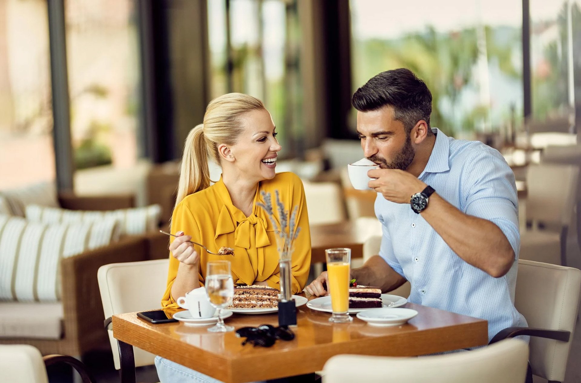 Smiling couple enjoying coffee and cake at a cafe table with a bright, modern interior.