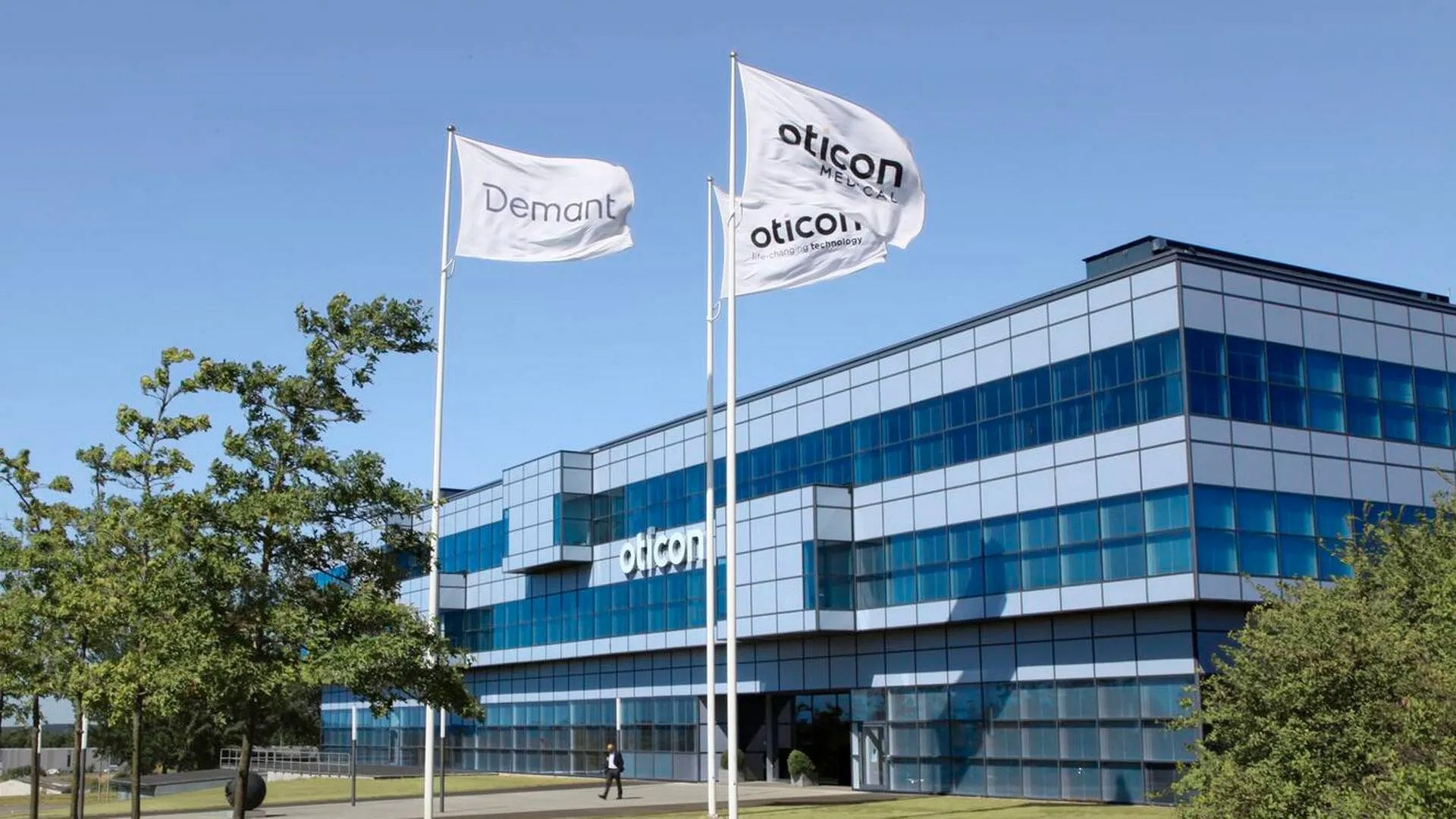 Modern glass office building with Oticon and Demant flags flying outside on a clear day.