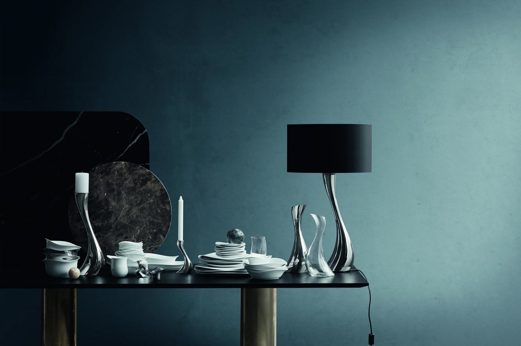 Artistic table arrangement with sculptural silver candlesticks, dishes, and a modern lamp against a dark blue wall.