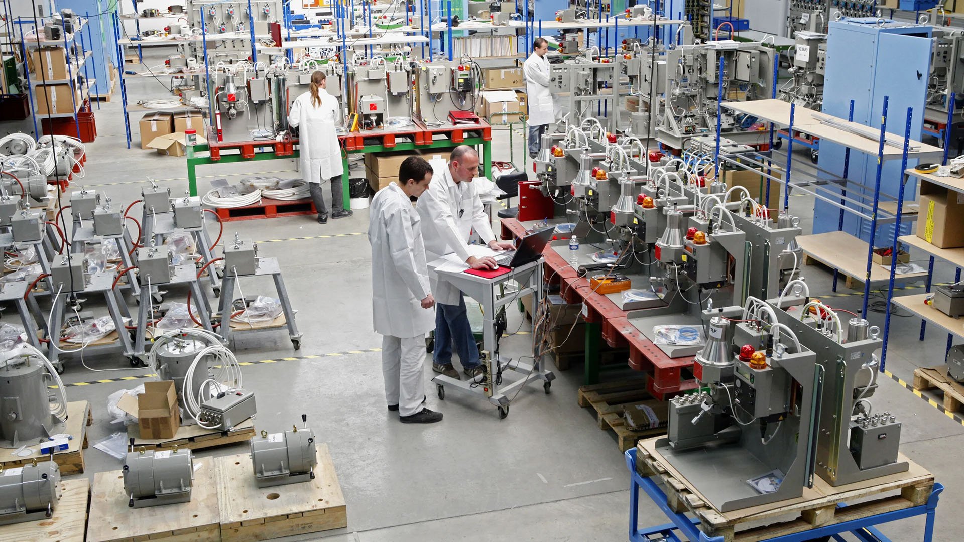 A busy manufacturing facility featuring workers in lab coats, various machines, and assembly stations with electronic equipment and parts.