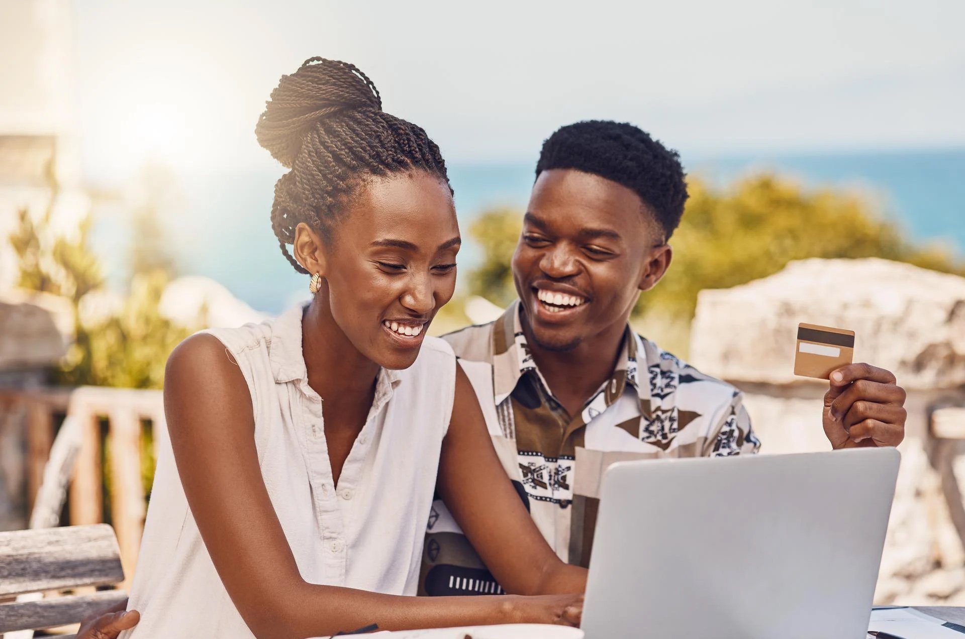 Smiling couple shopping online with a laptop and credit card outdoors on a sunny day.