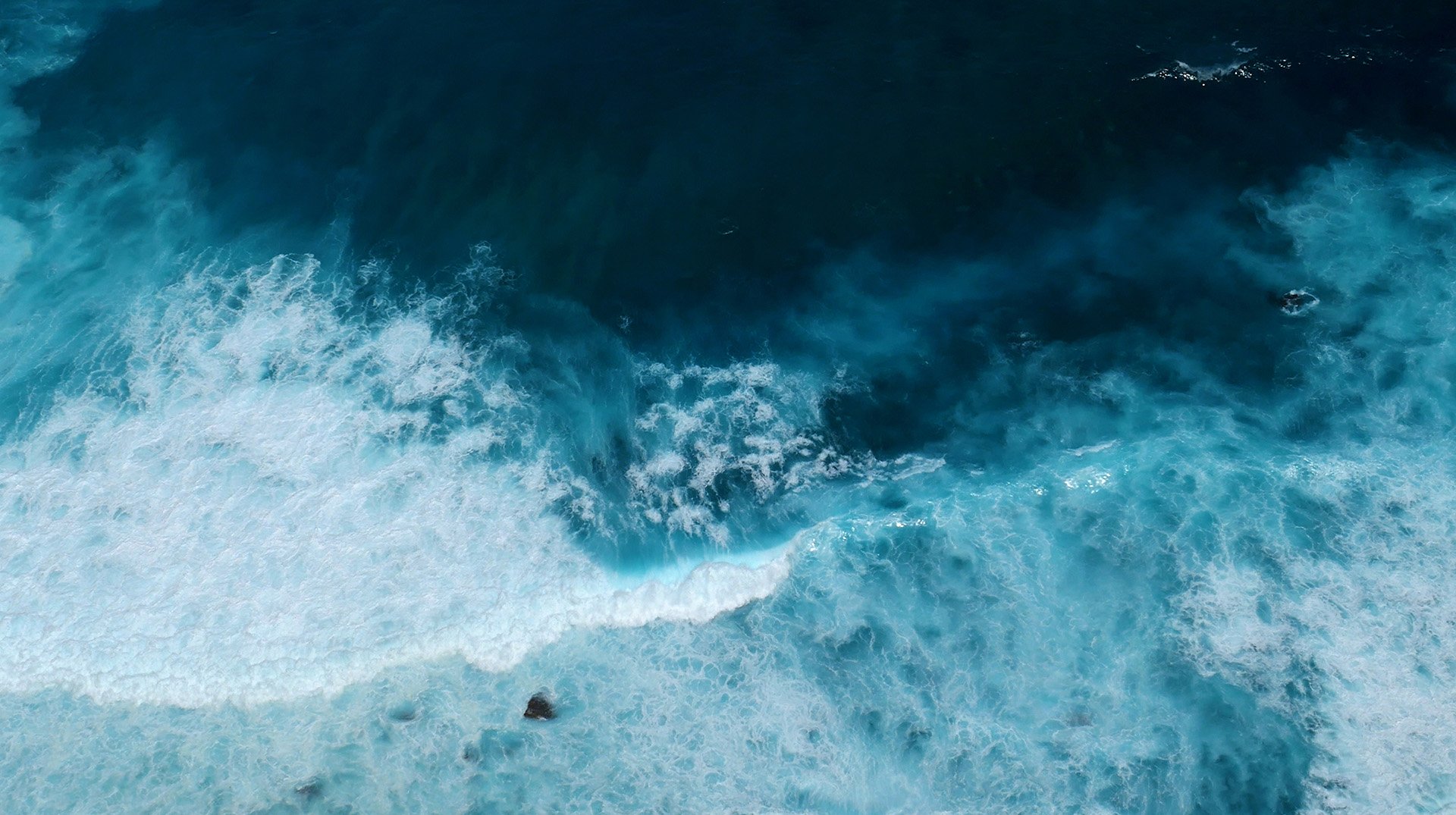 Aerial view of vibrant blue ocean waves crashing onto a rocky shore, creating white frothy foam against deep turquoise waters.