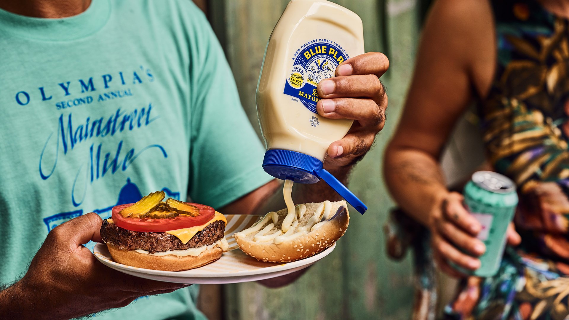 Person squeezing Blue Plate mayonnaise onto a hamburger bun at an outdoor gathering.