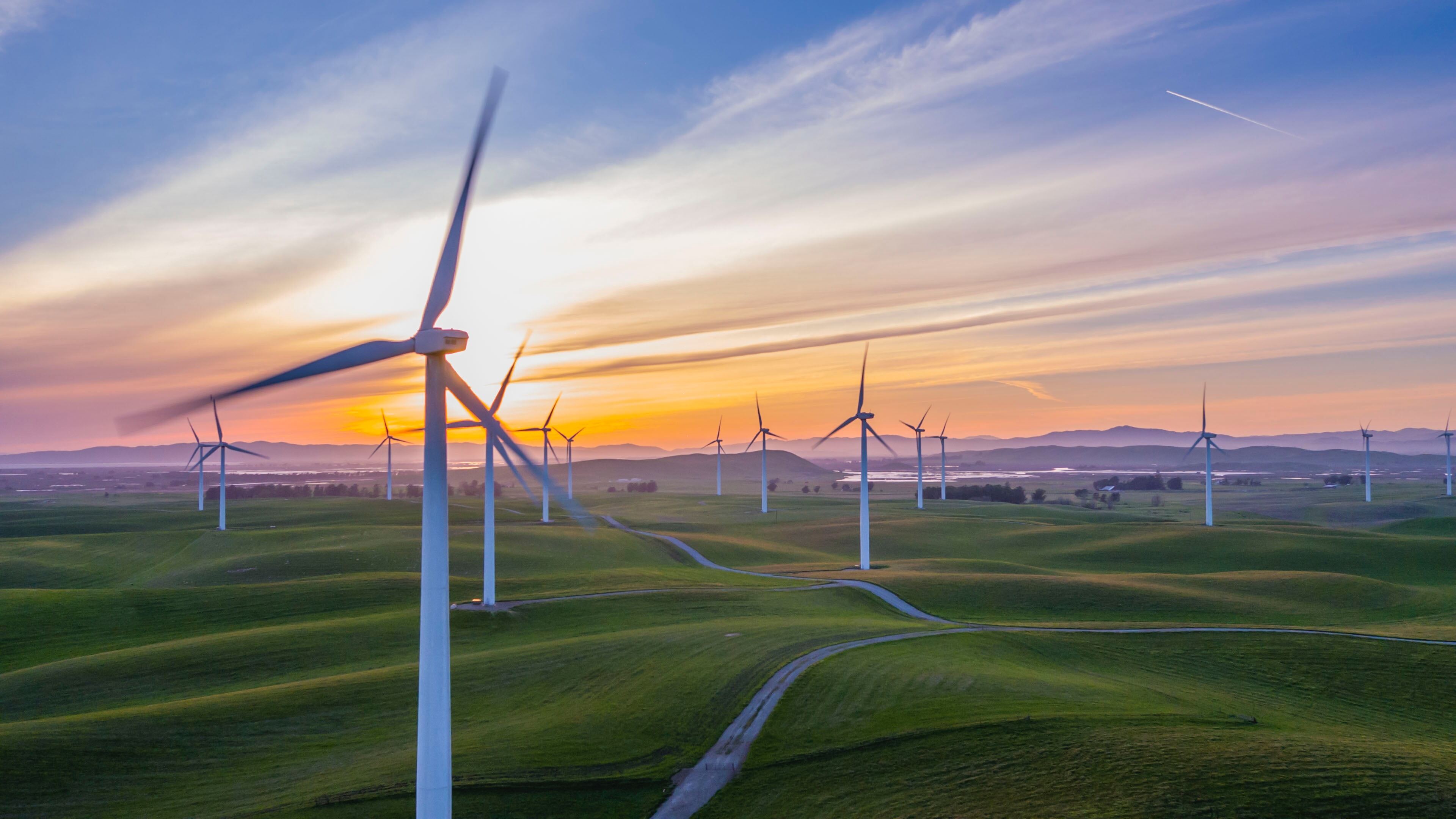Wind turbines on a green landscape during sunset with colorful sky in the background.