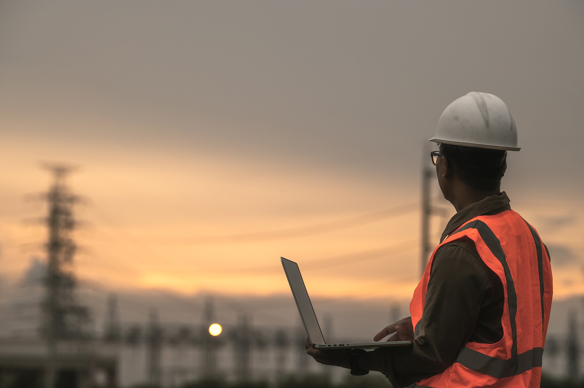 A construction worker in a hard hat and orange vest observes a sunset near power lines, holding a laptop. The scene conveys focus and contemplation.