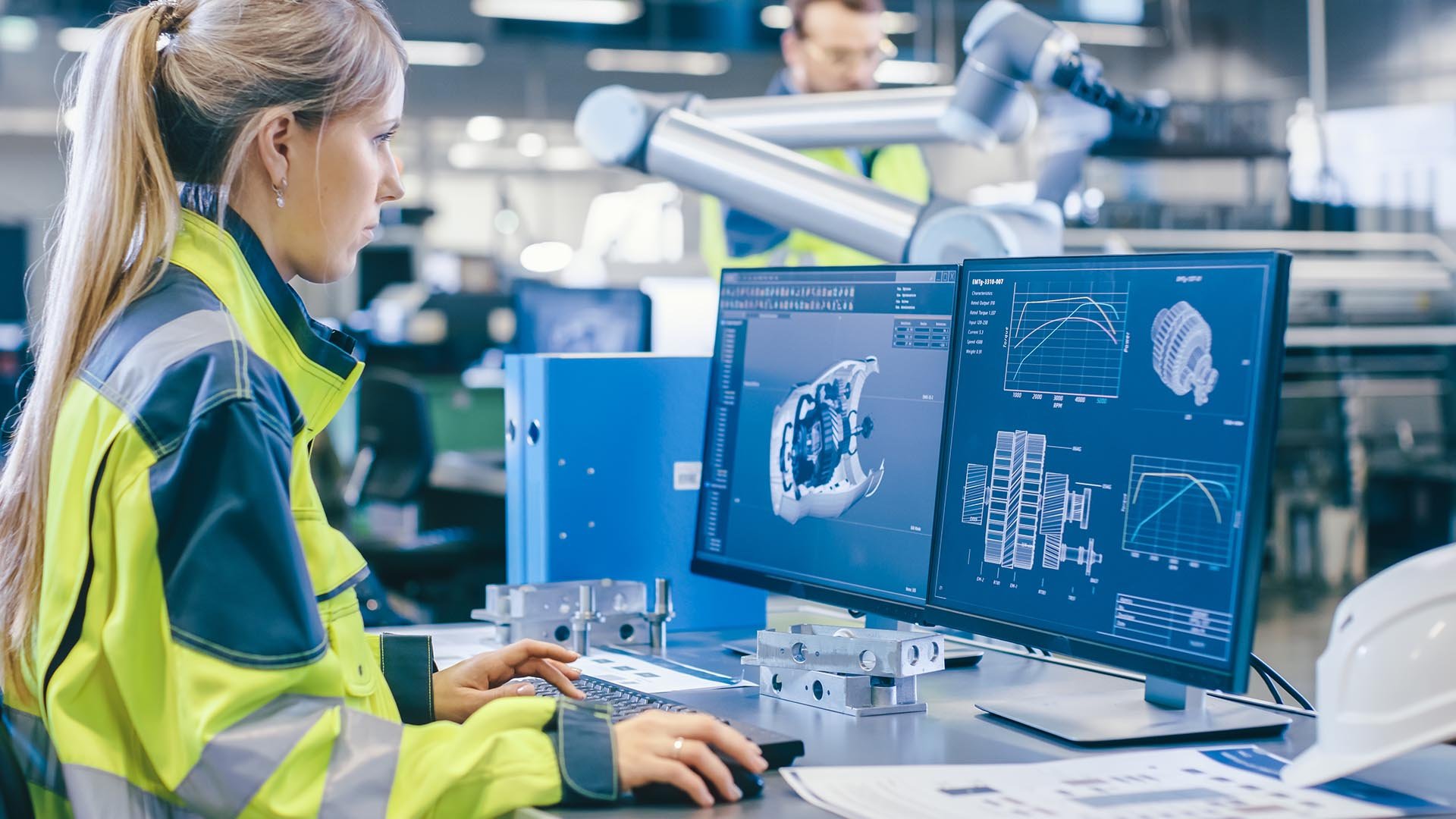 Une personne vêtue d'une veste de sécurité lumineuse travaille sur un ordinateur affichant des schémas techniques dans un environnement industriel moderne.