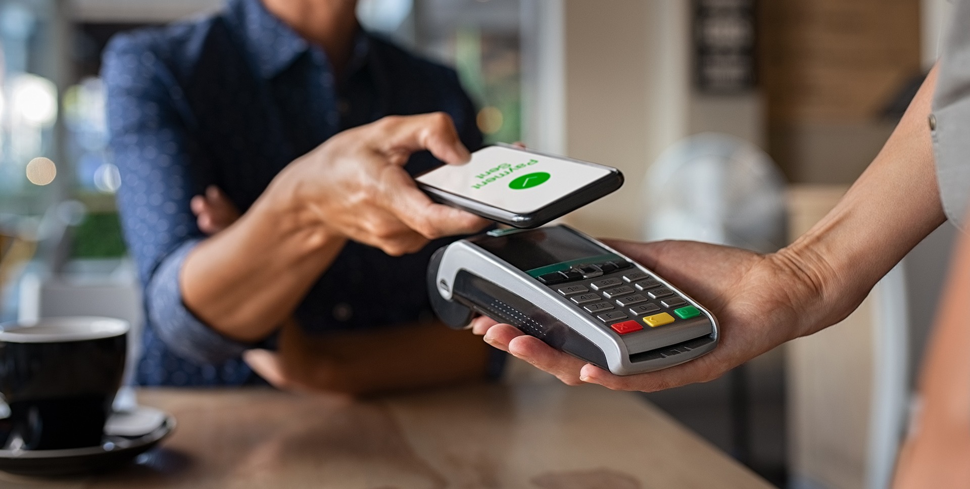Smiling person makes a contactless payment with a smartphone over a card reader in a cafe. A coffee cup is visible on the table.