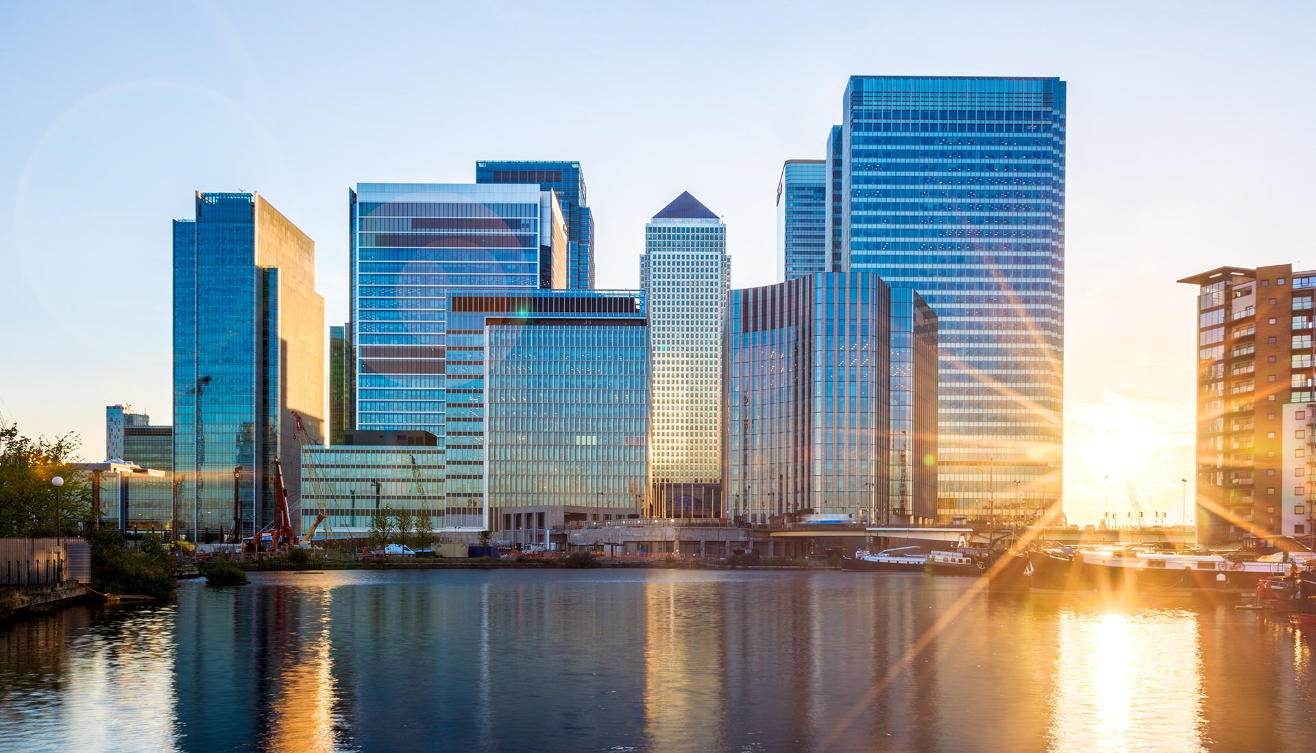 Sunset over a city skyline with modern glass skyscrapers reflecting the golden light. A calm river is in the foreground, enhancing the serene atmosphere.