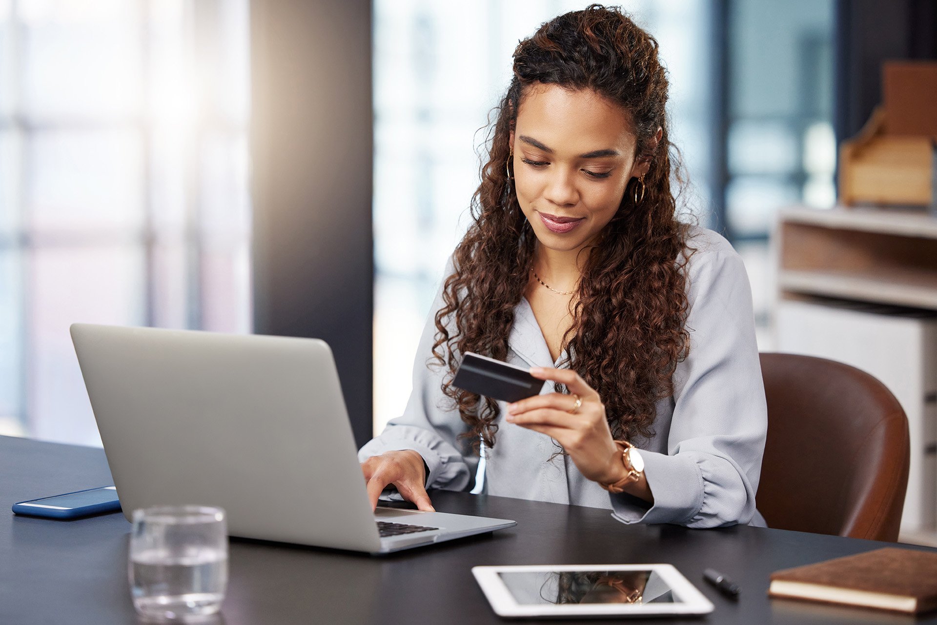 Woman with curly hair sits at a modern desk, holding a bank card and using a laptop. A tablet, phone, and glass of water are nearby. Bright, focused mood.