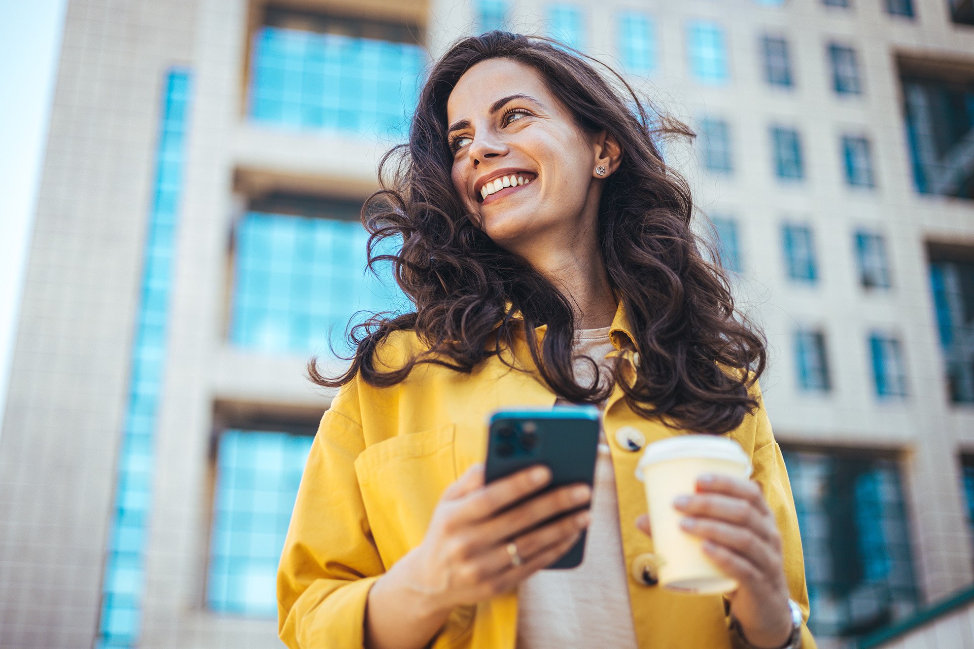 A woman in a yellow jacket holds a smartphone and a coffee cup, standing in front of a modern building with large glass windows.