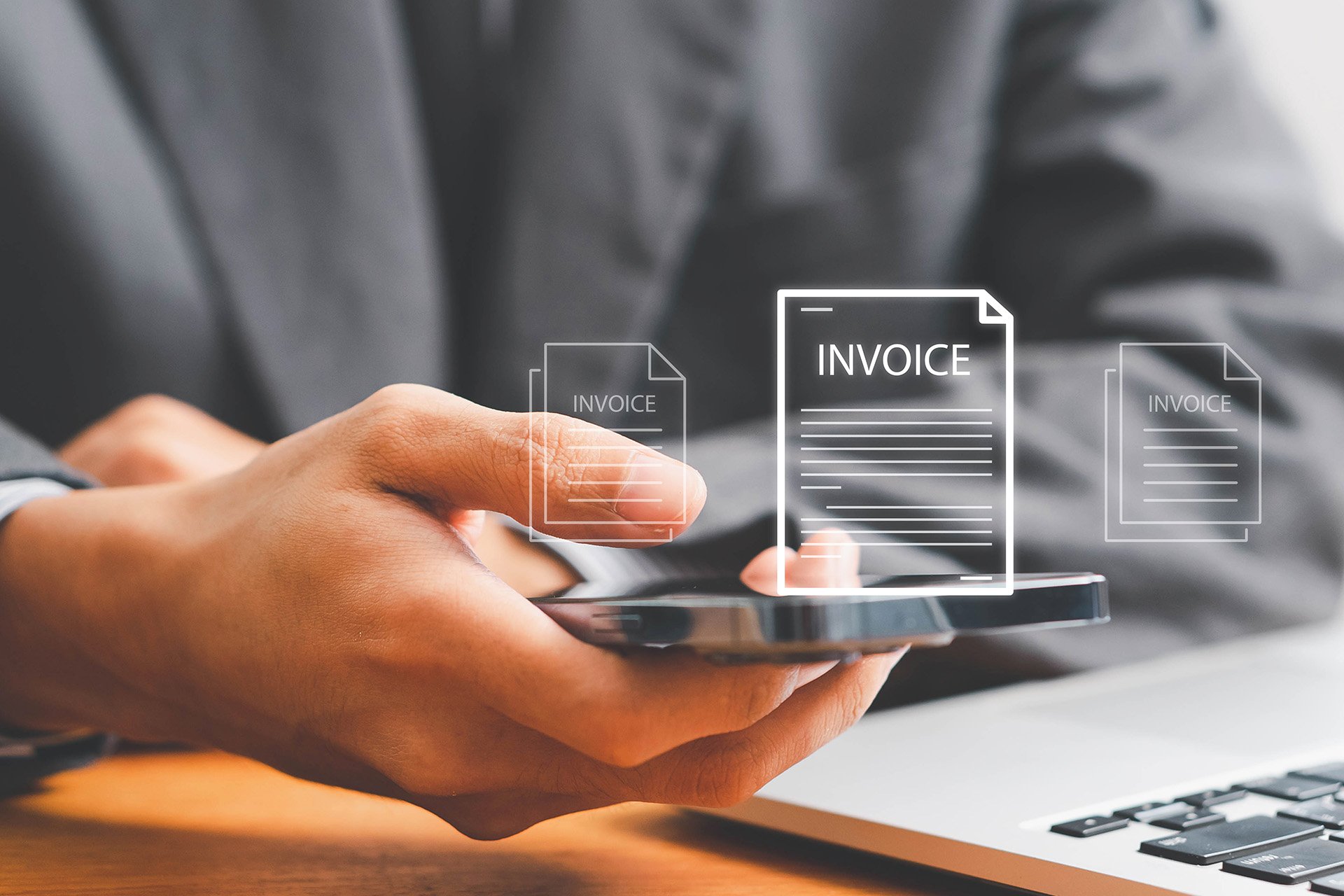 A person in a suit holds a smartphone displaying floating invoice icons while seated at a desk with a laptop.