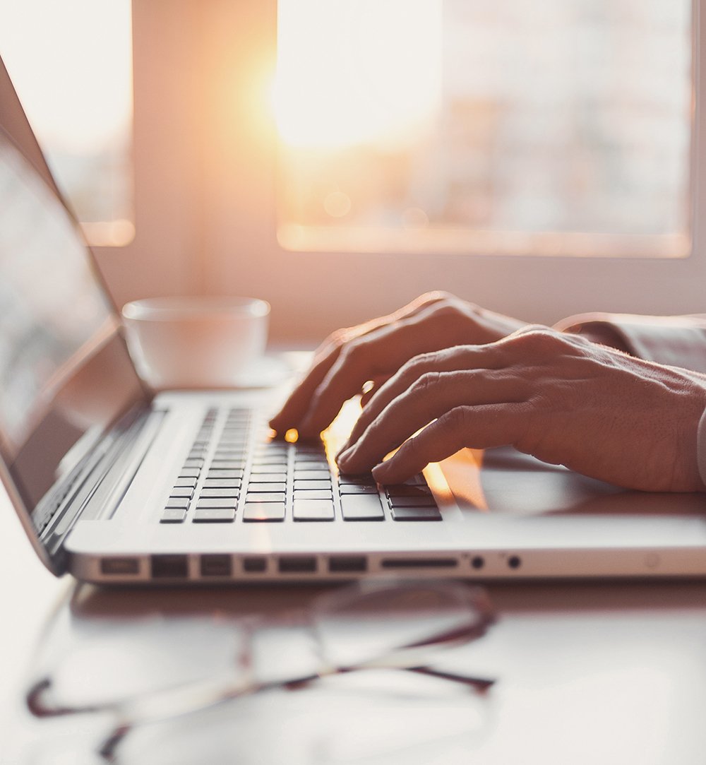 A person's hands typing on a laptop, with a warm sunset glow and a coffee cup in the background on a well-lit table.