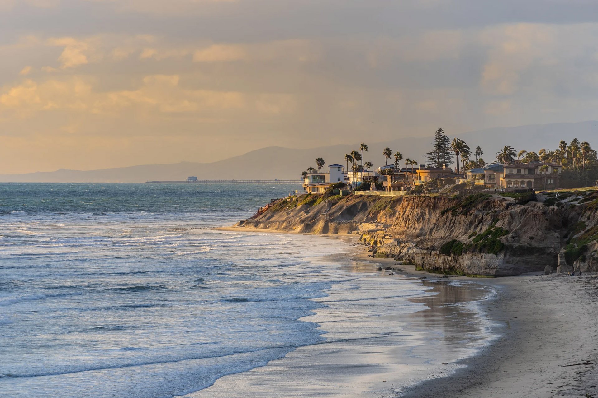Coastal view of a sandy beach with homes on cliffs, palm trees swaying, and gentle waves under a golden sunset sky.