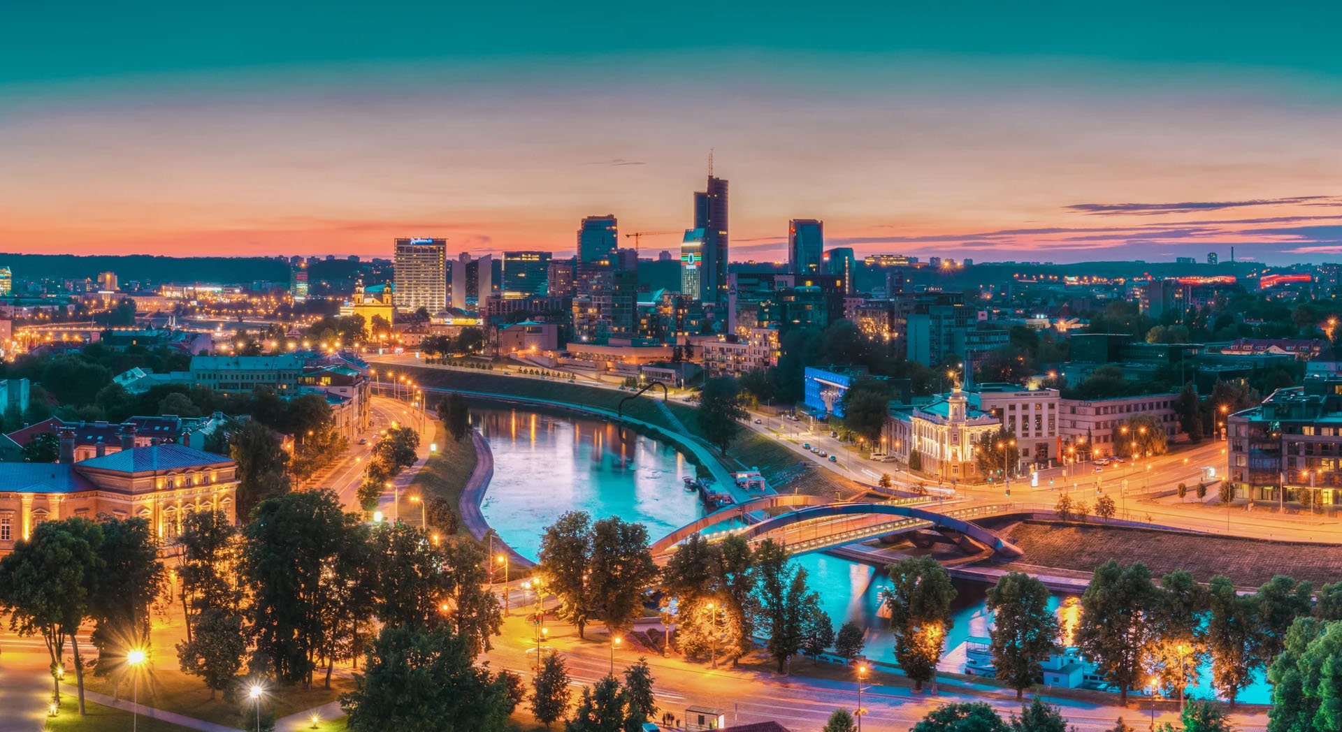 Panoramic view of a vibrant cityscape at sunset, featuring illuminated buildings, a winding river, and a bridge.