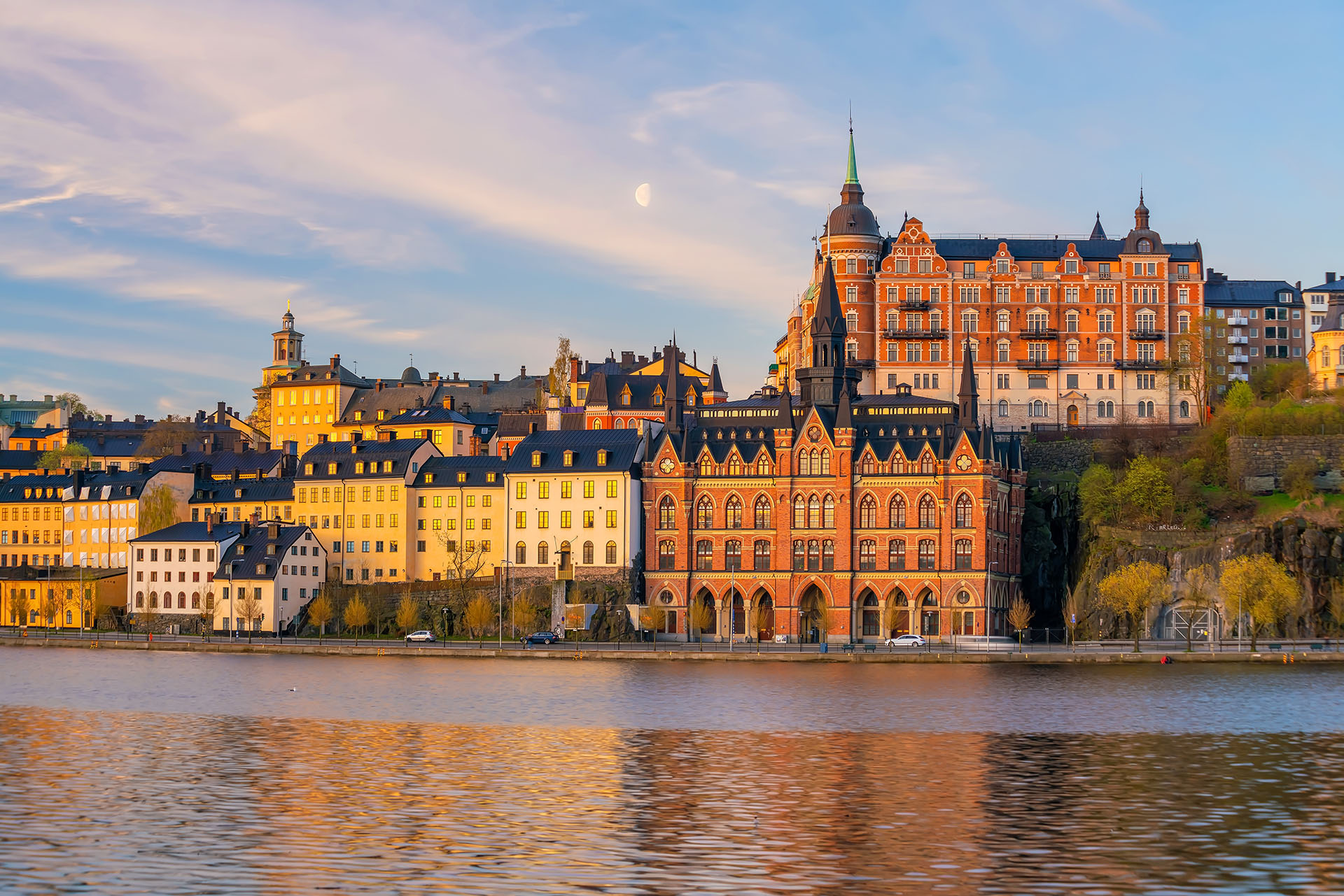 Golden hour light illuminates historic buildings along the water in Stockholm, with a crescent moon visible in the sky.
