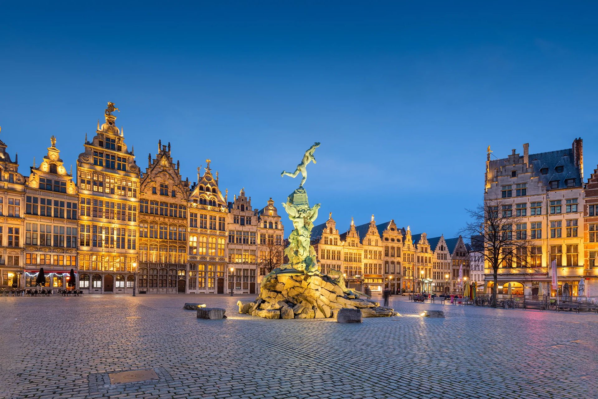 Historic square at dusk with ornate, illuminated buildings and a central statue on a rocky base, creating a tranquil, majestic atmosphere.