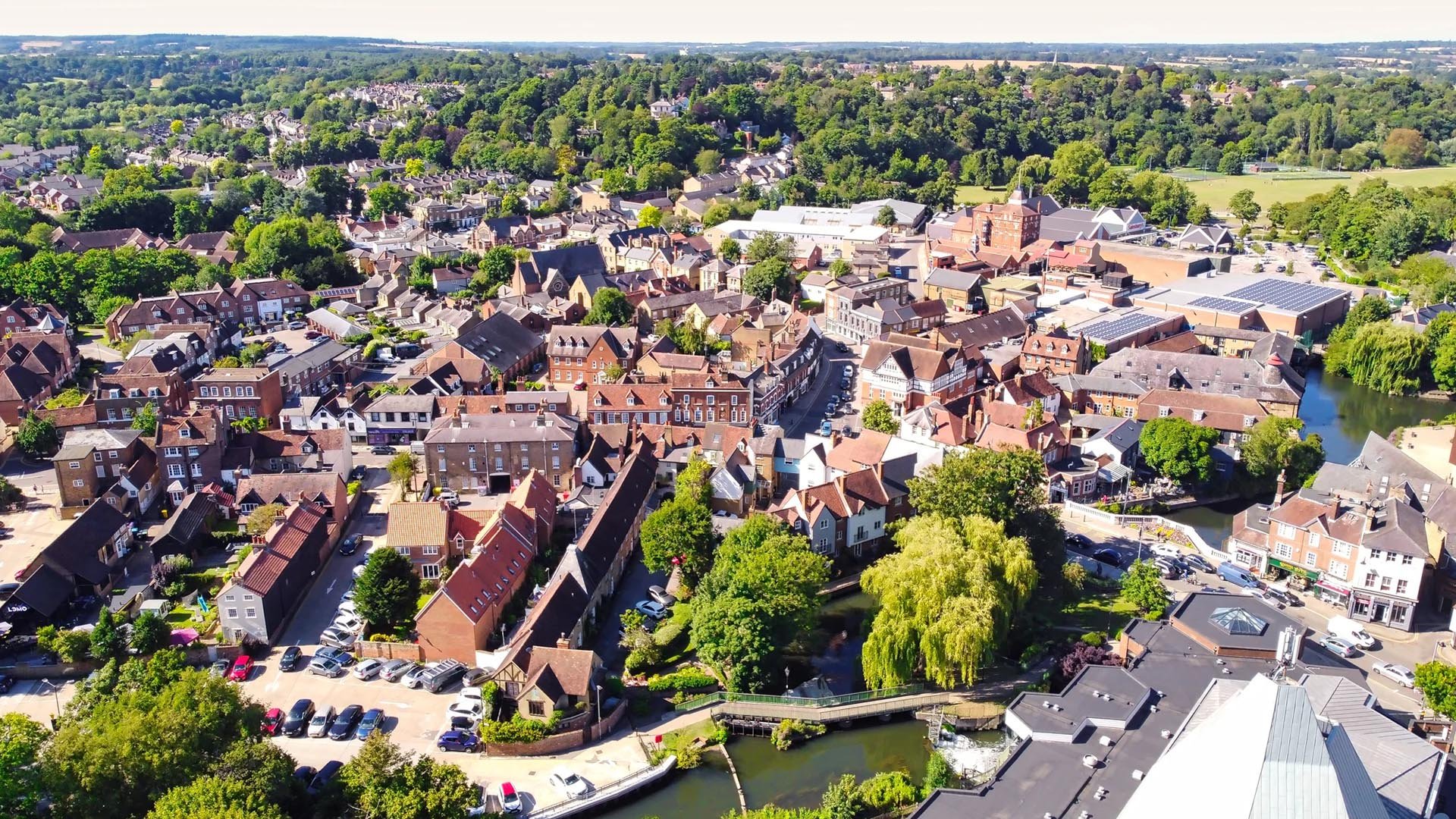 Aerial view of a quaint English town with red-roofed buildings, narrow winding streets, lush green trees, and a serene river flowing through.