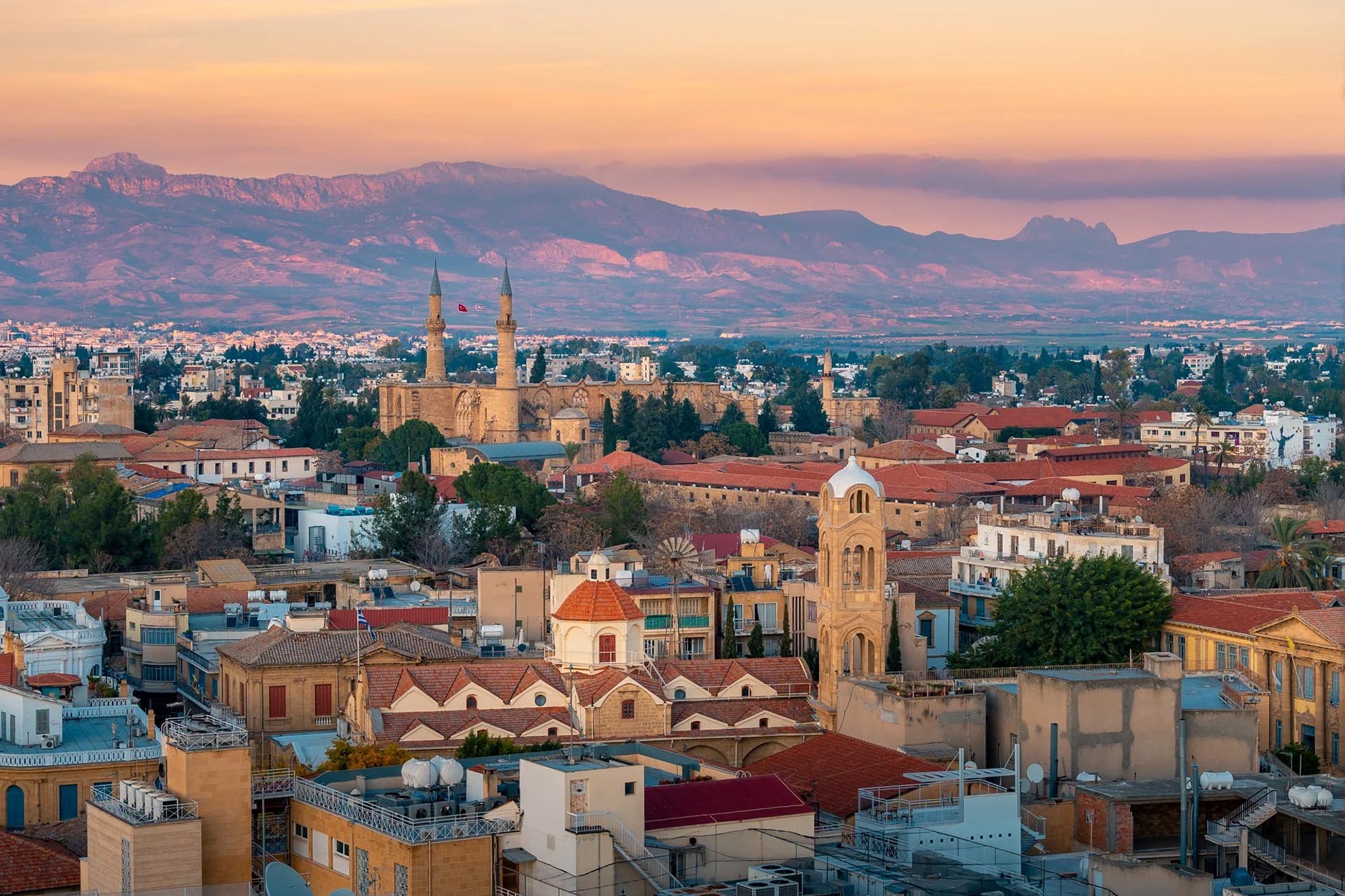 Panoramic view of a cityscape at sunset with a blend of historic and modern architecture. Red-roofed buildings and a mosque, with distant mountains.