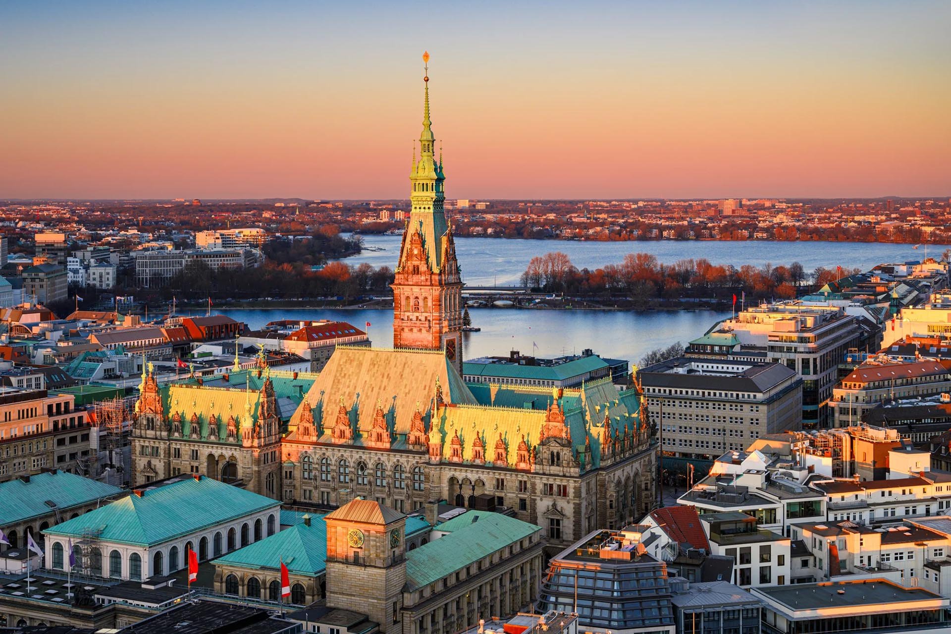 Aerial view of Hamburg's city hall at sunset, featuring a tall spire, intricate architecture, and a serene lake in the background, under a pastel sky.