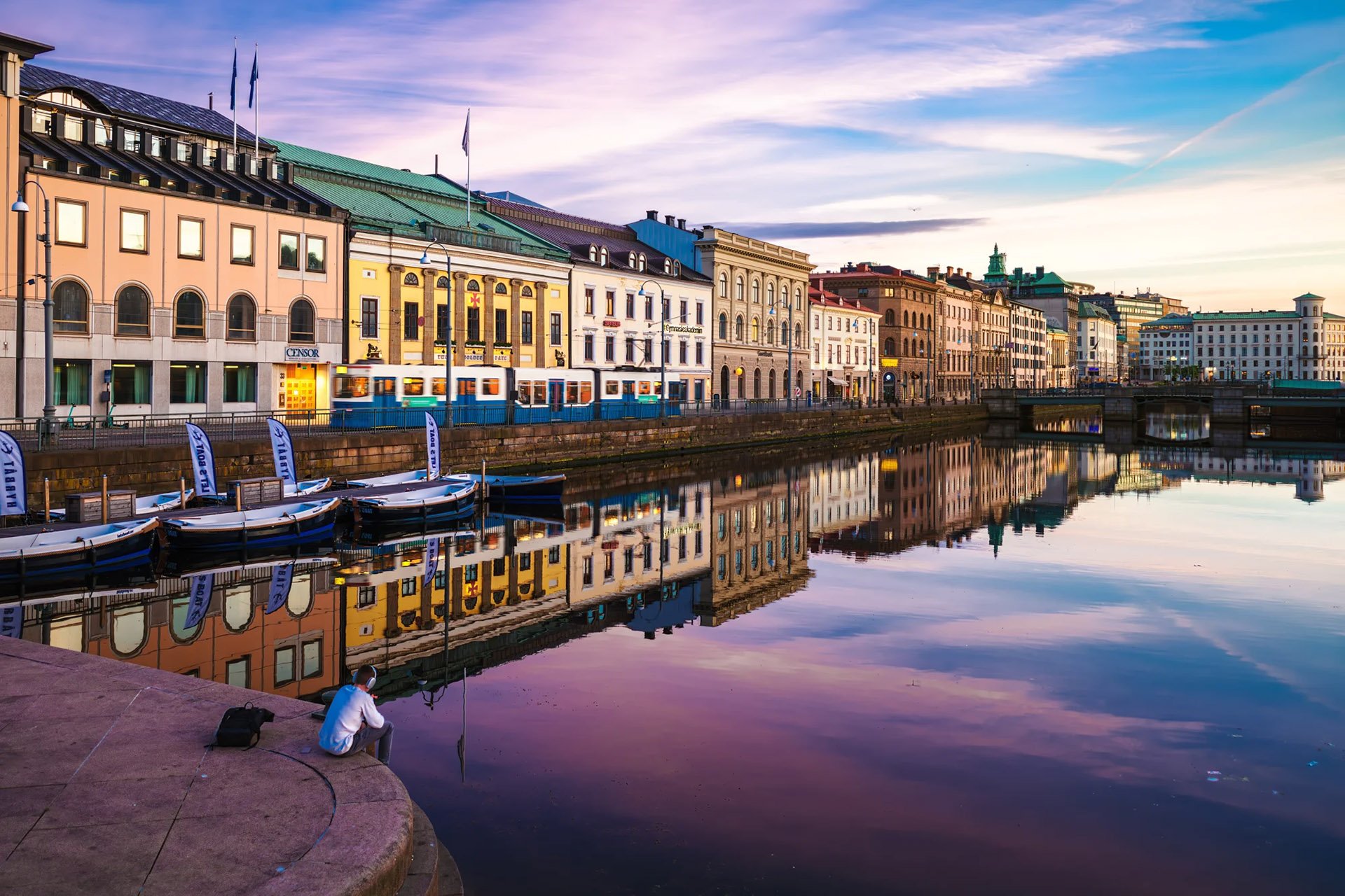 A tranquil waterfront scene at sunset with colorful buildings reflected in calm water. A person sits pensively on a riverbank near docked boats.