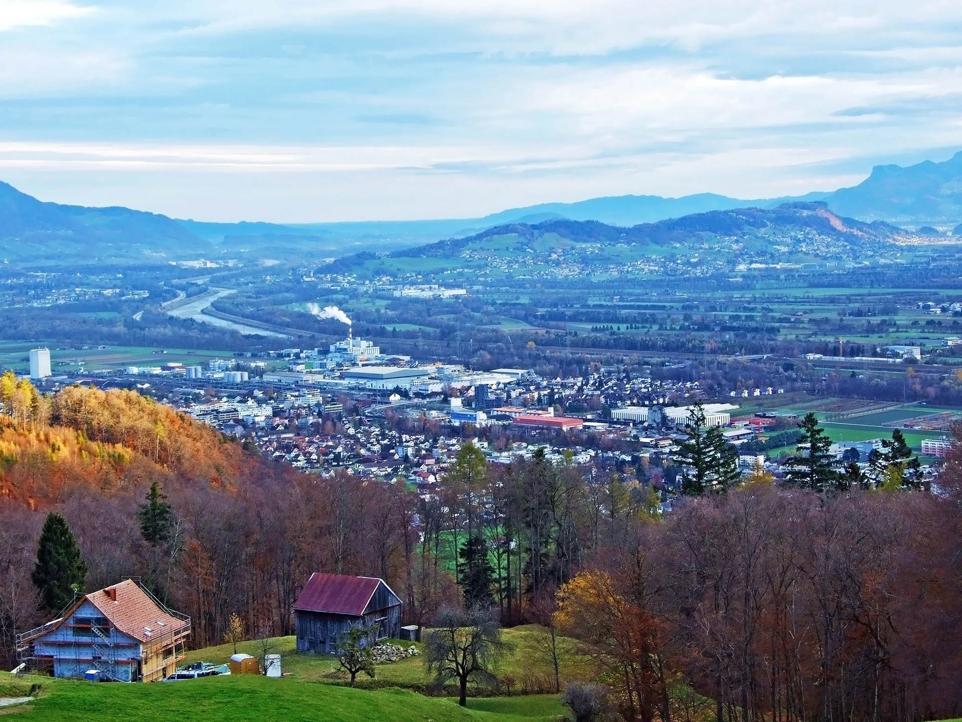 Expansive landscape view of a quaint town nestled in a valley, surrounded by autumn trees. Rolling hills and distant mountains under a cloudy sky.