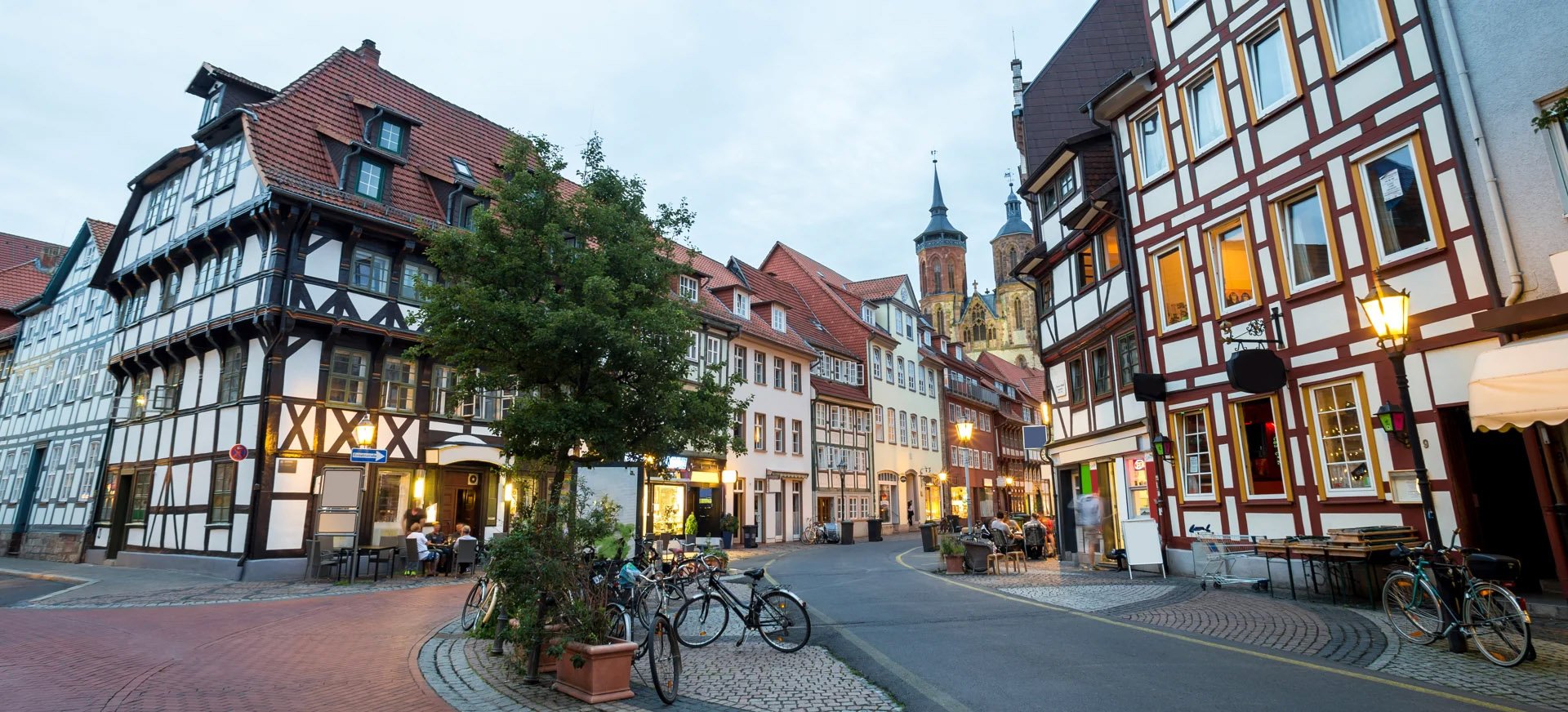 Historic street scene with charming half-timbered buildings, a church spire, and bicycles parked near a tree. Warm lights give a cozy evening ambiance.