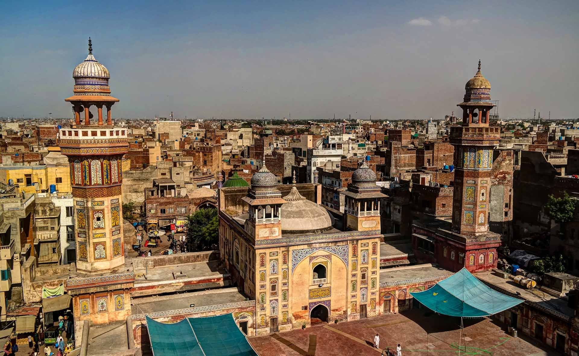 Aerial view of the historical Wazir Khan Mosque in Lahore, Pakistan, with intricate minarets and domes. The bustling old cityscape surrounds it.