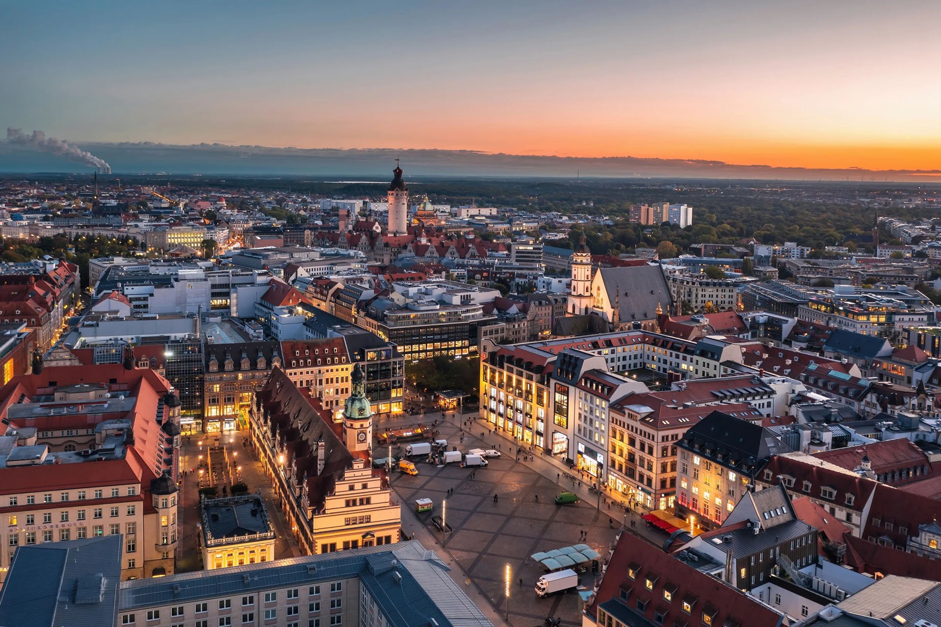 Panoramic view of a city at dusk, showcasing illuminated historic buildings, a clock tower, and a central square against a colorful sunset sky.