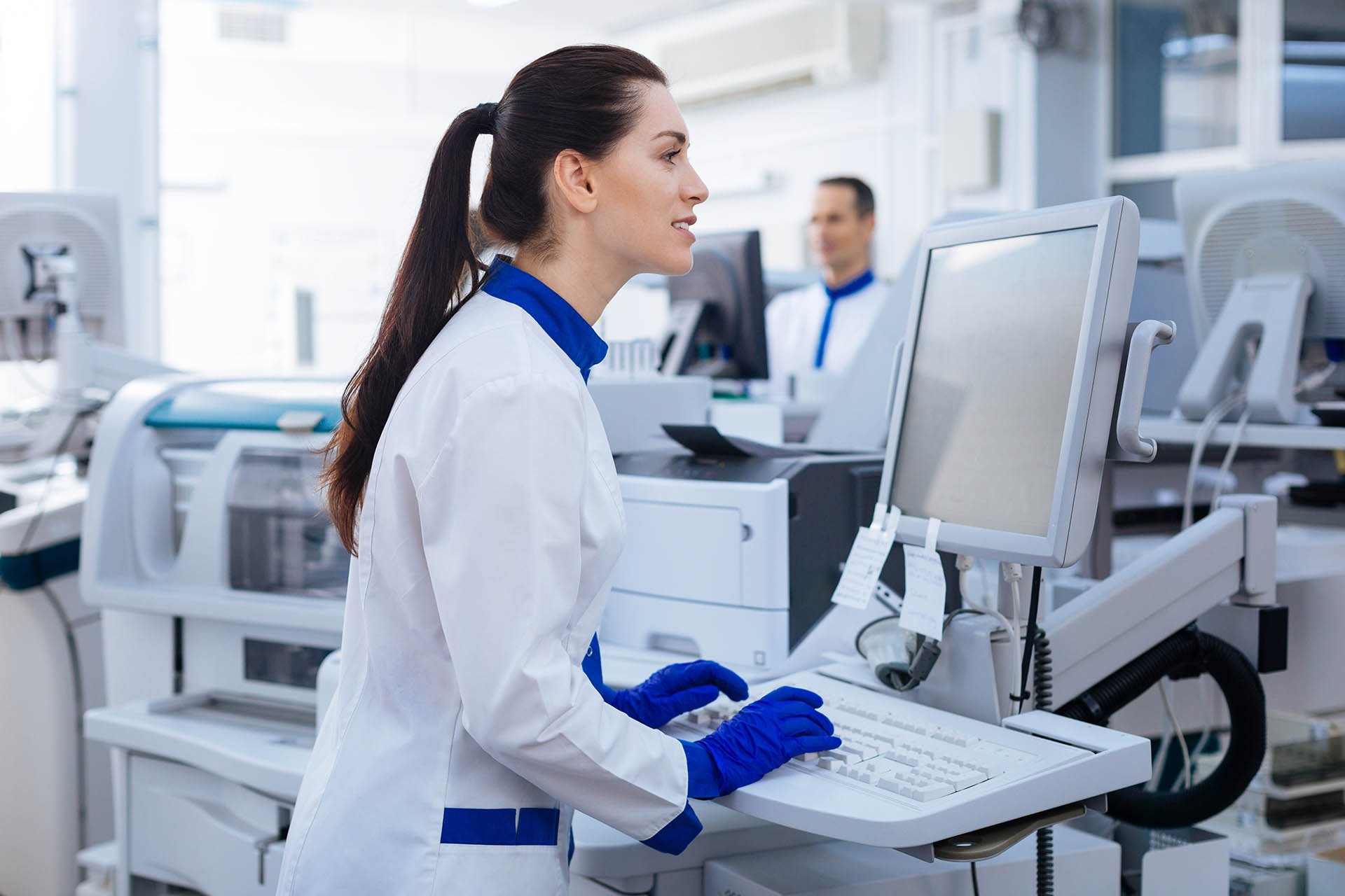 A laboratory technician in a white coat and blue gloves works at a computer, surrounded by medical equipment in a bright workspace.
