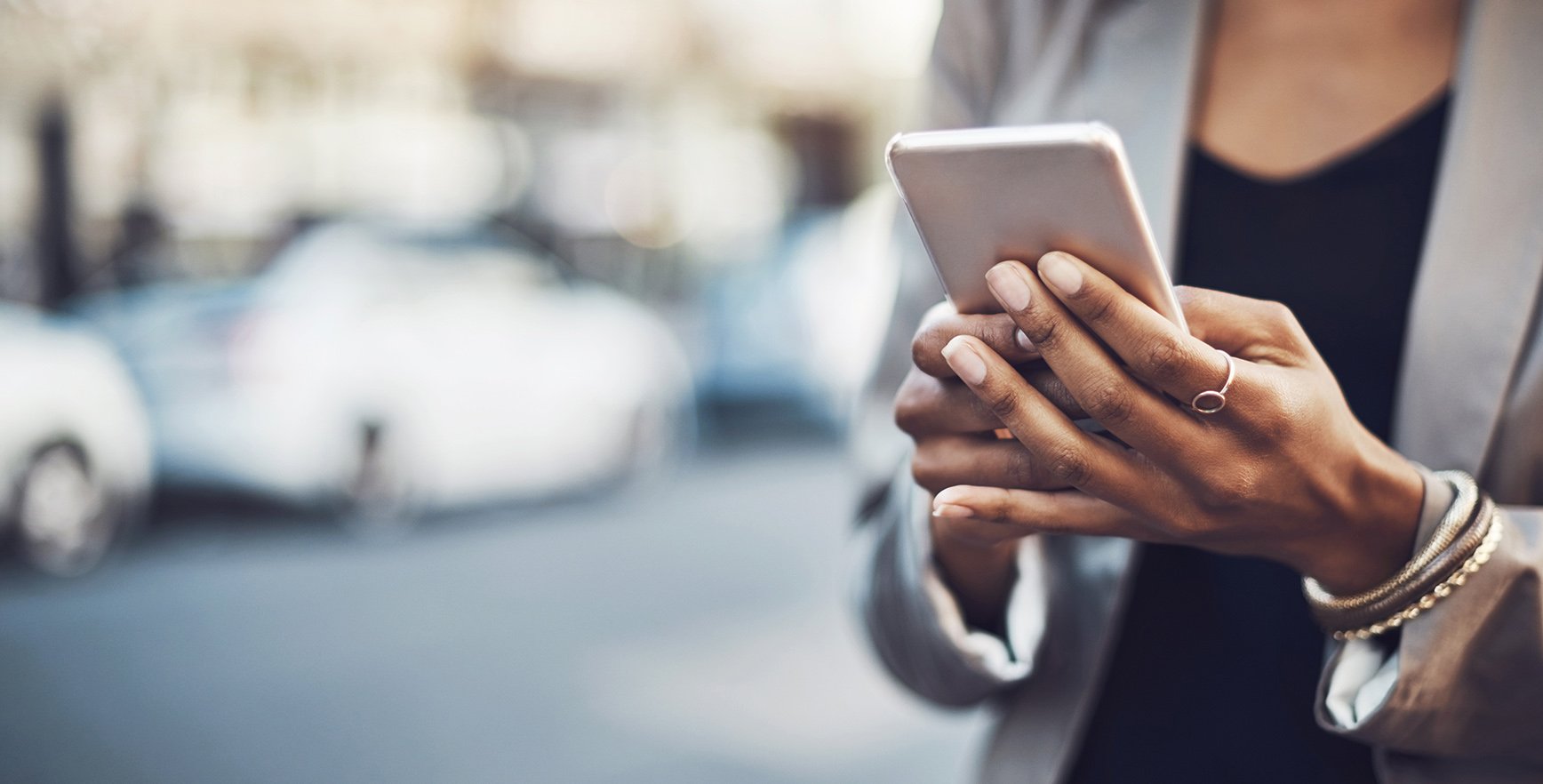 A close-up of a person's hands holding a smartphone, with blurred cars in the background, conveying a sense of urban life.