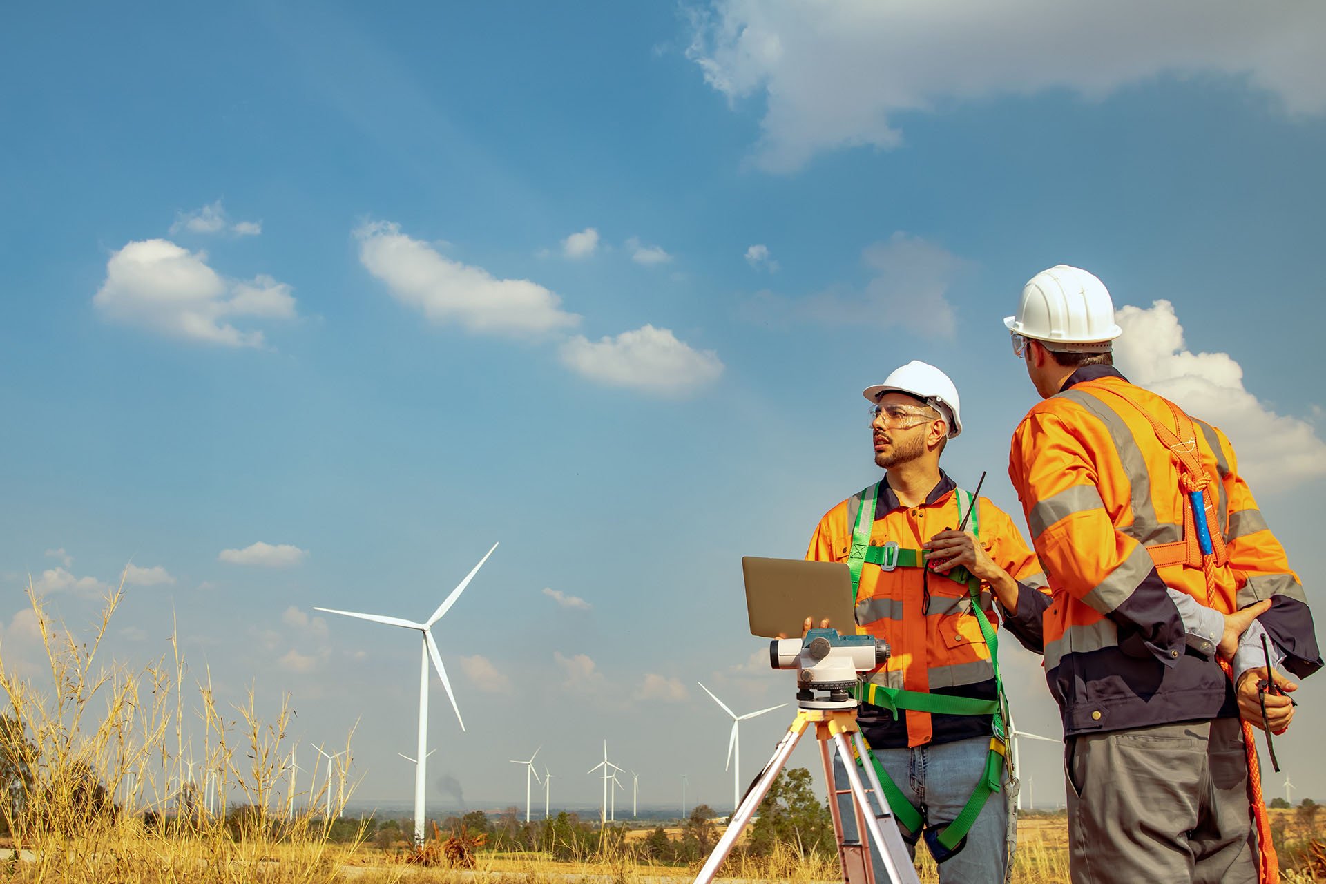 Two men in orange vests stand beside large wind turbines against a clear blue sky.