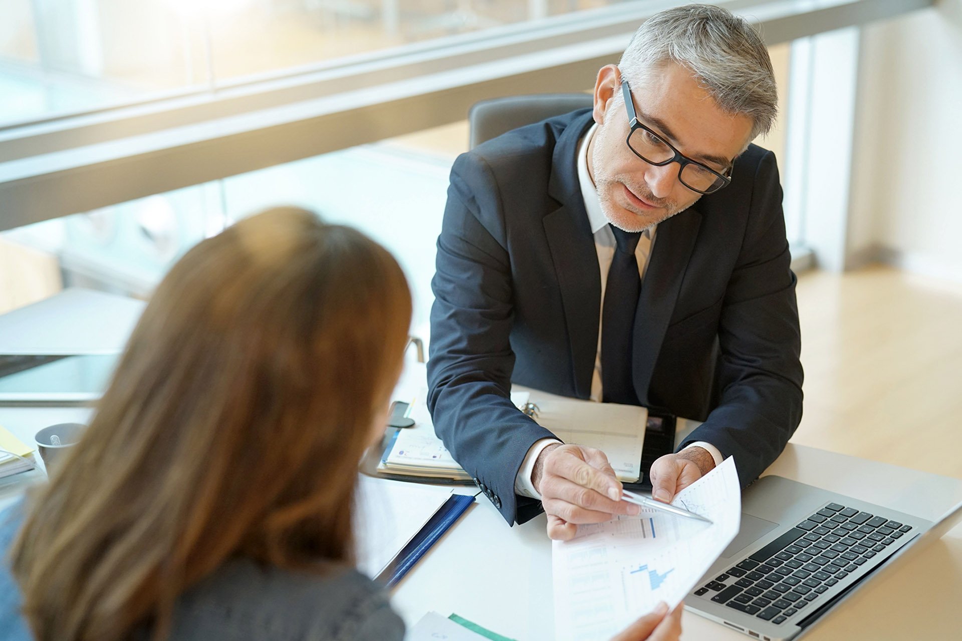 A business meeting taking place at a modern office, with documents and a laptop on the table, as two people discuss financial reports.