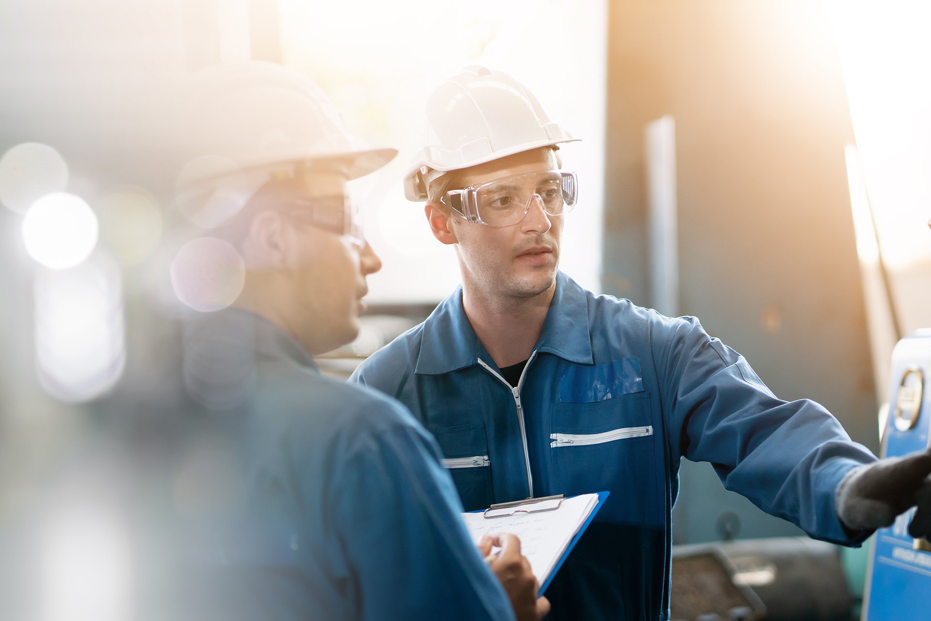 Two engineers wearing blue uniforms, hard hats, and safety glasses are discussing while reviewing a clipboard. The setting is a bright, industrial environment.
