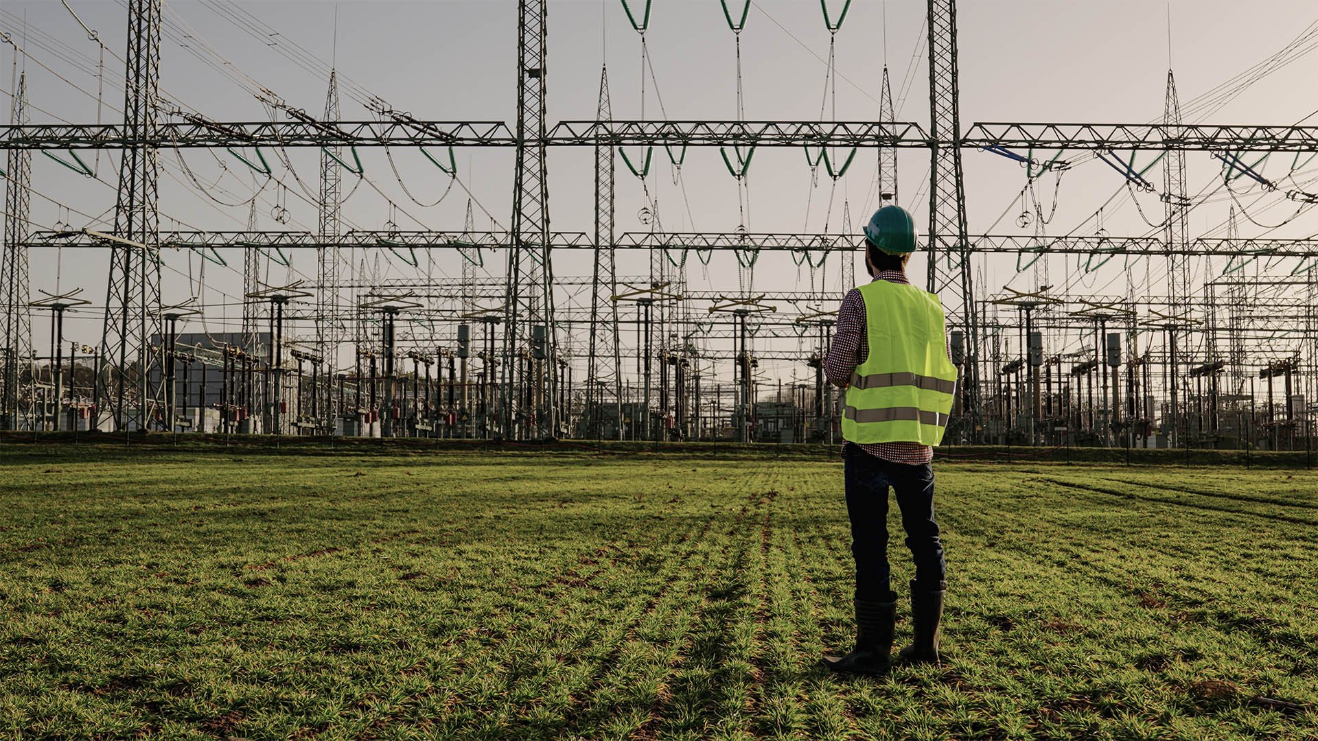 A man in a yellow vest stands in front of an electrical substation, overseeing operations and safety measures.